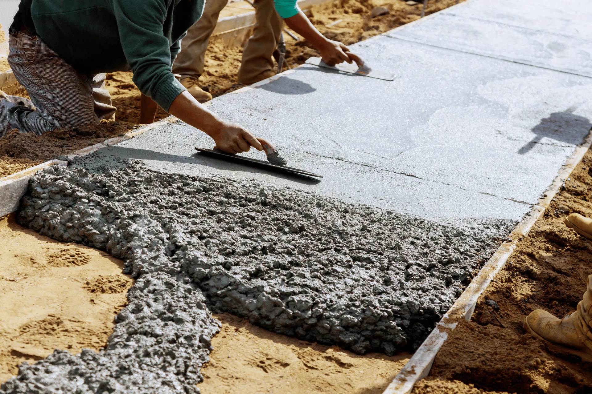 Construction workers are pouring and smoothing wet concrete to form a sidewalk. The image shows the process of building a new walkway with freshly mixed concrete being worked by hand with trowels. Several workers are visible, with their hands and tools actively engaged in shaping the surface of the concrete.