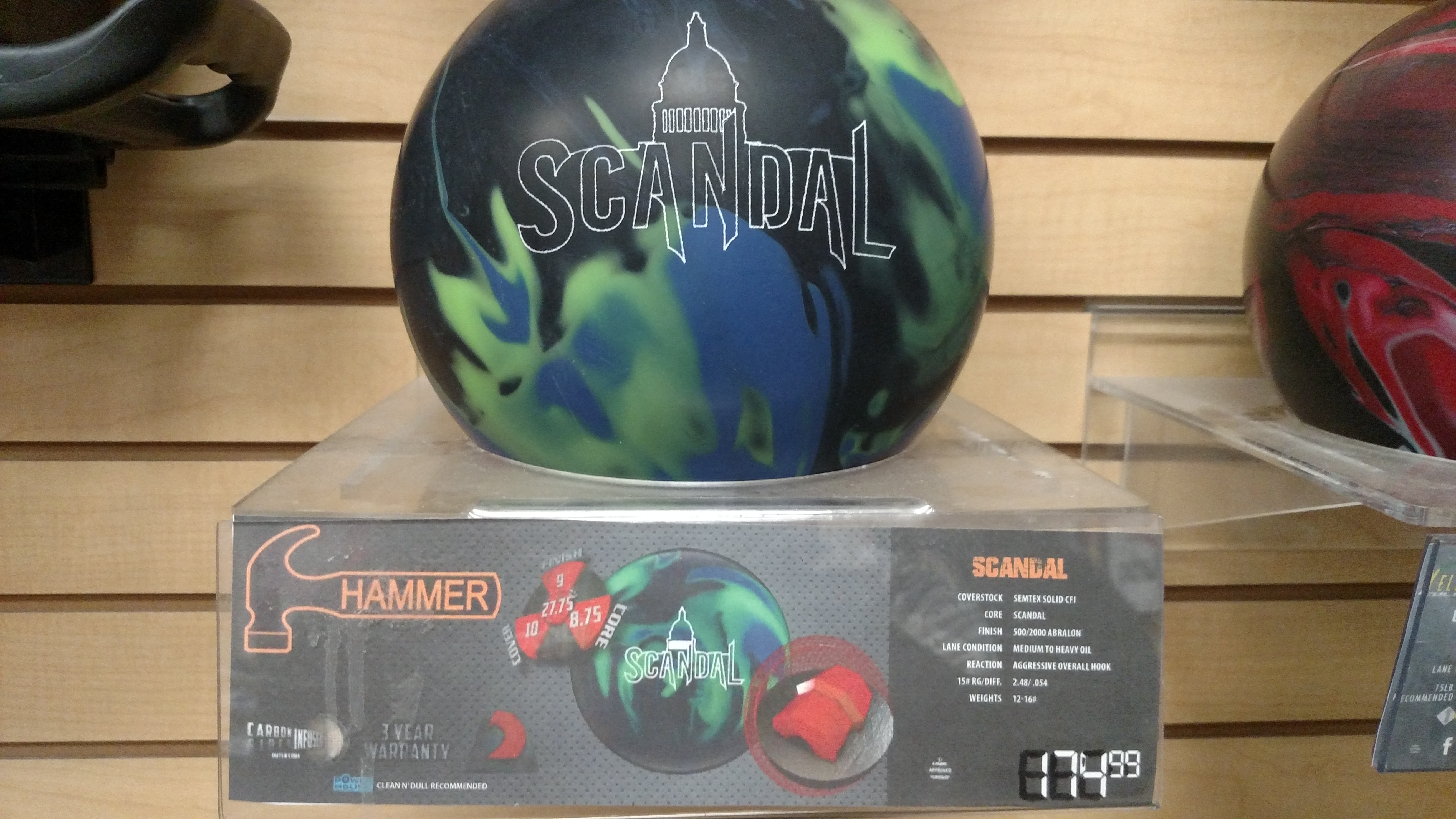 A close-up shot of a bowling ball with a black, green, and blue marbled design, featuring the brand name "SCANDAL" in white lettering with a silhouette of a dome building above it. The ball is displayed on a clear acrylic stand, which also shows a label with product details and pricing. To the right, part of another bowling ball, predominantly red and black, is visible. The background consists of light brown wooden panels.