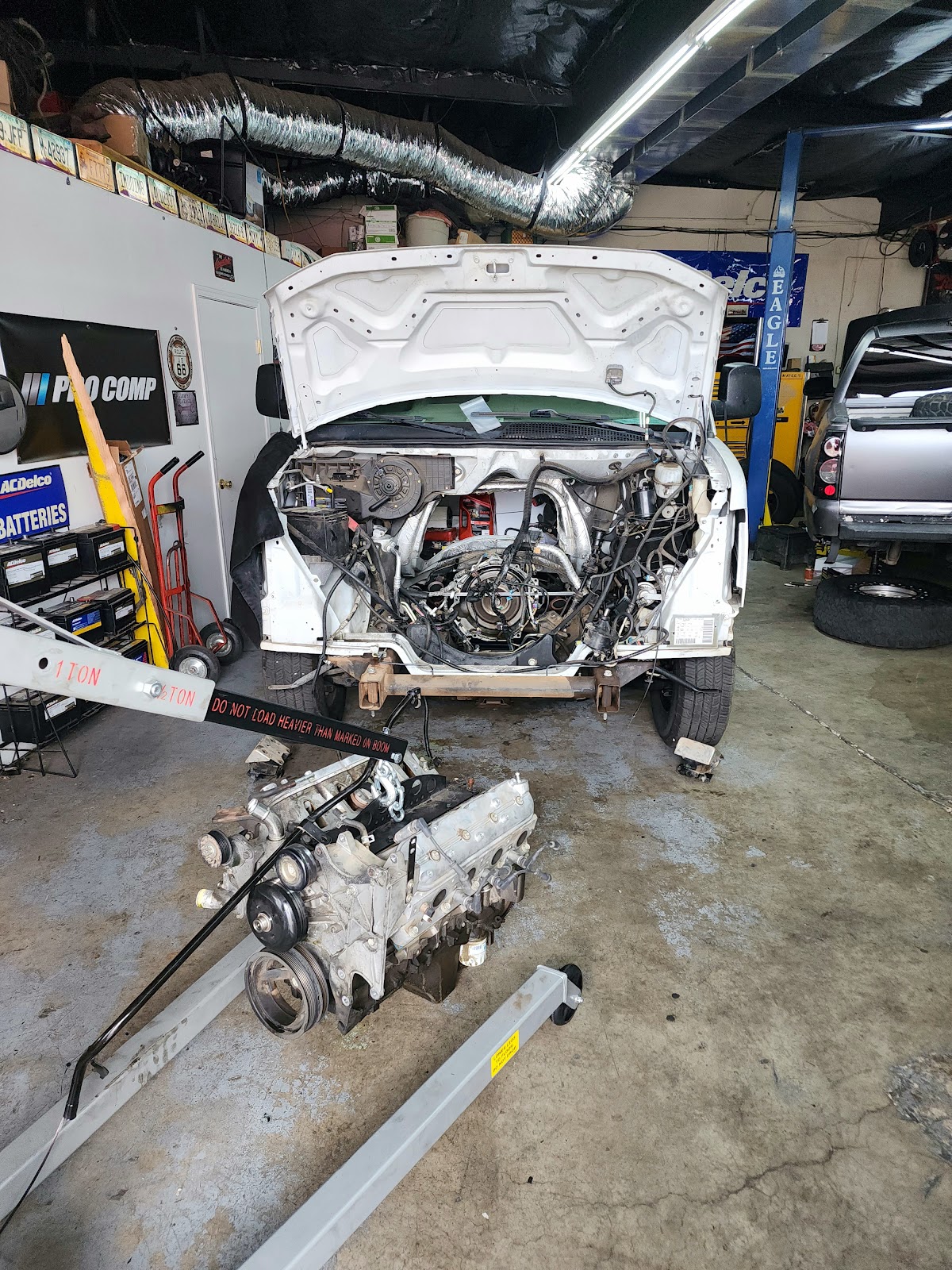 A white van is being worked on in a garage. The engine has been removed and is sitting on an engine hoist in front of the van. The front of the van is open, exposing the engine bay and various wires and hoses. Another vehicle, a silver truck, is parked to the right of the van. The garage appears to be a professional auto repair shop, with tools, equipment, and car parts visible throughout.
