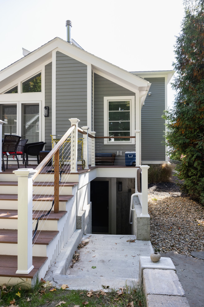A side view of a modern gray house with a large wooden deck and a staircase leading down to a basement entrance. The deck features white railings with horizontal cable supports and wooden steps. The house has a gabled roof with several windows, including a triangular one. To the right of the house, a dense green tree stands tall, and the ground is covered with gravel and some scattered leaves. The overall scene suggests a residential property, possibly during autumn due to the fallen leaves.