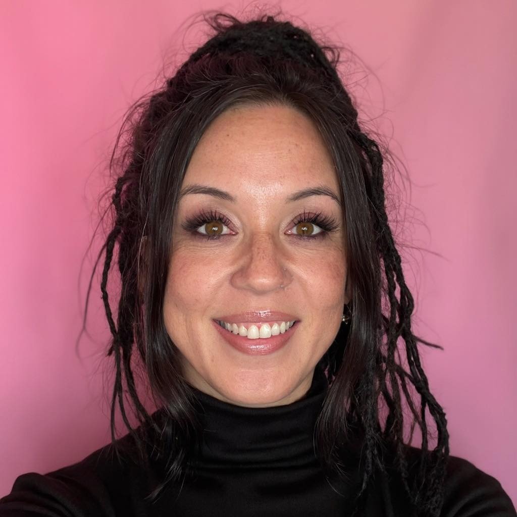 Professional headshot of Rowena Tulacz, President and Founder of R. Construction Solutions. Rowena is photographed against a warm pink background wearing a classic black turtleneck. She has dark hair worn in locs styled upward, warm brown eyes, and a confident, approachable smile. The portrait conveys both professionalism and warmth &mdash; reflecting her 30+ years of hands-on construction industry expertise and her reputation as a trusted partner and advisor to contractors nationwide.