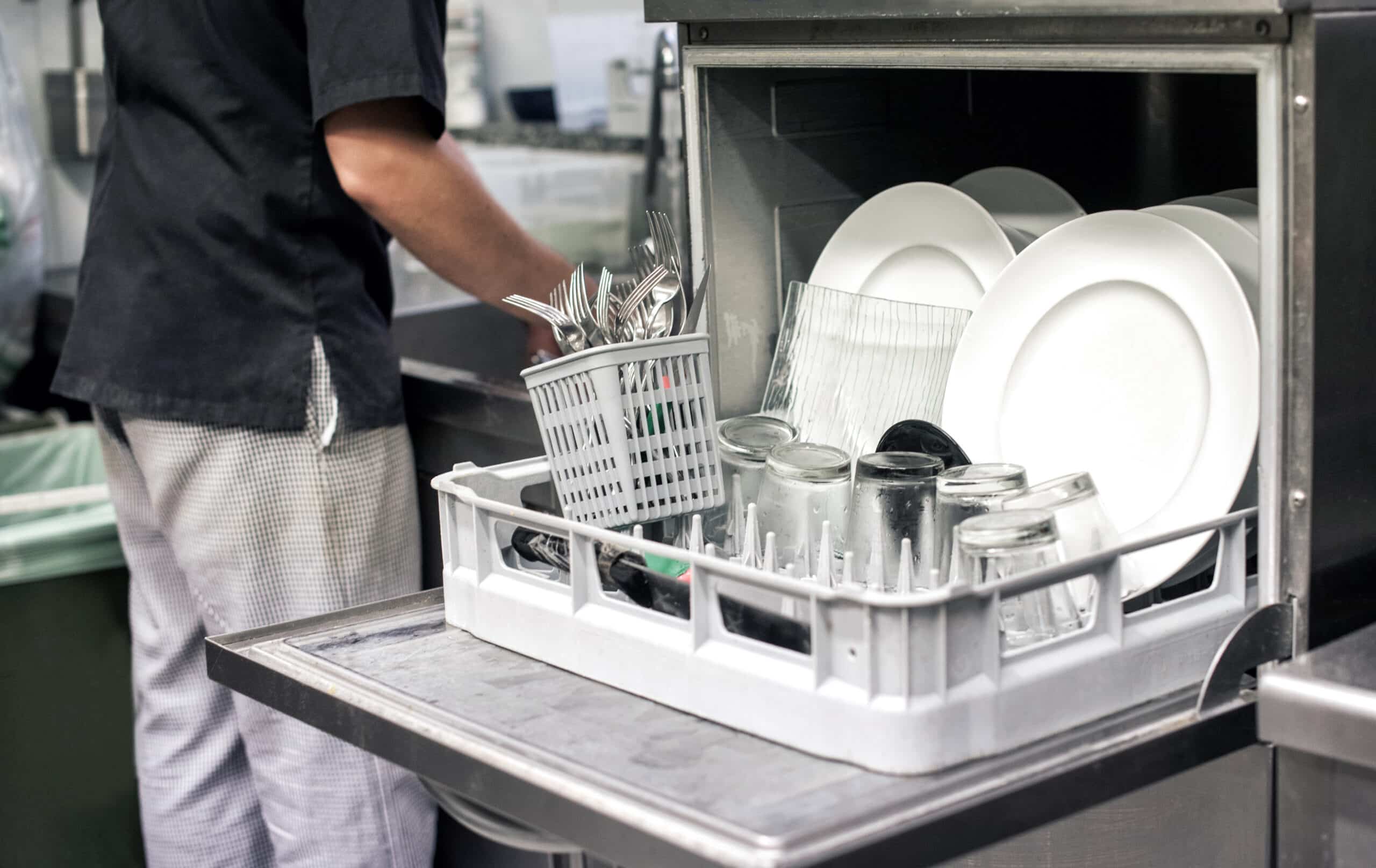 Kitchen hand with an open dishwasher filled with clean white plates in a restaurant kitchen in a catering and hygiene concept