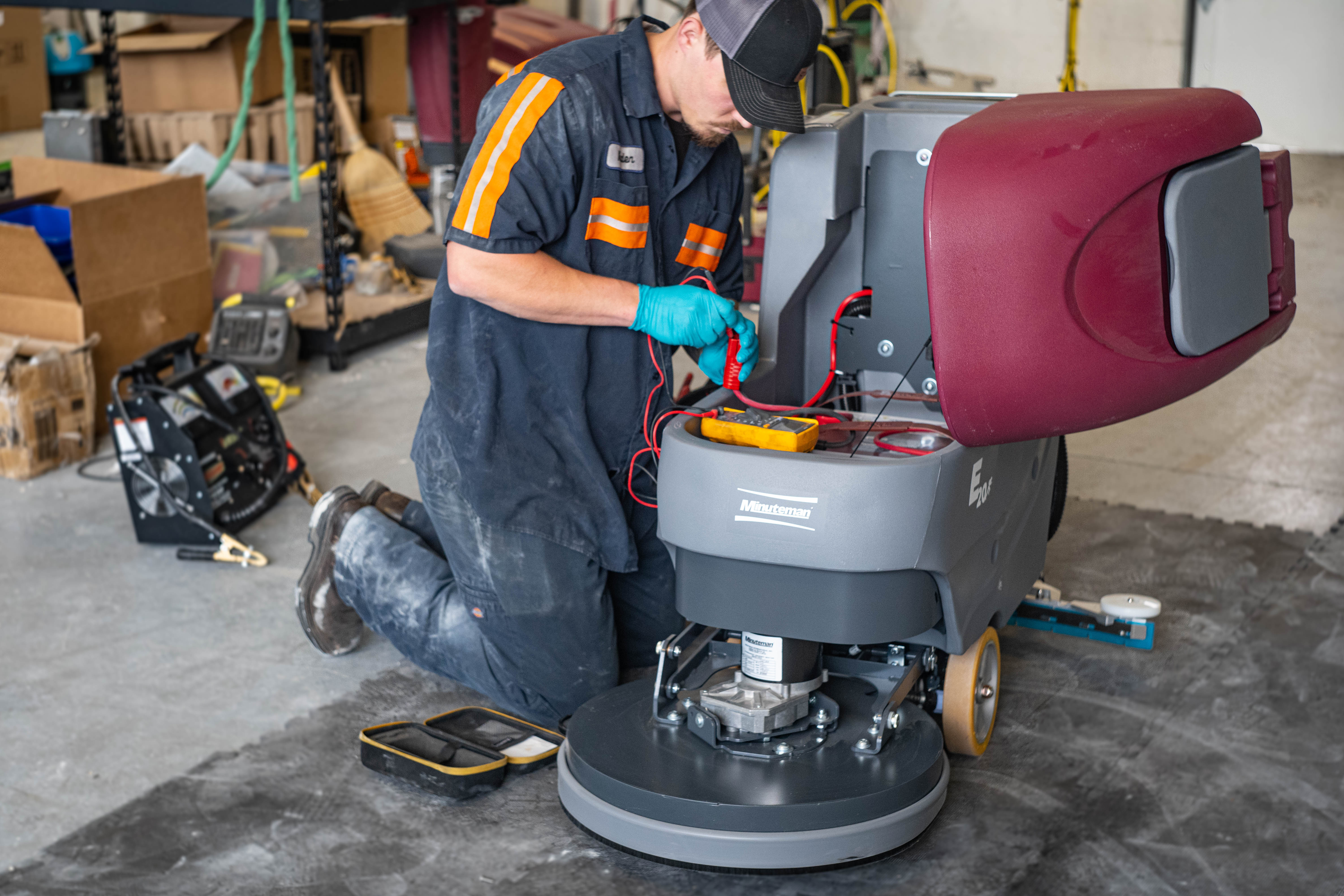 A technician kneeling next to an open Minuteman E20 floor scrubber unit, using a digital multimeter to test live electrical circuitry and internal wiring currents.