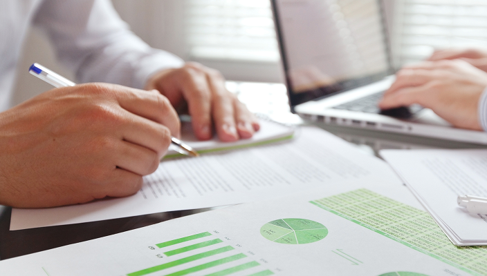 Close-up of two people working in an office with a laptop and spreadsheets.