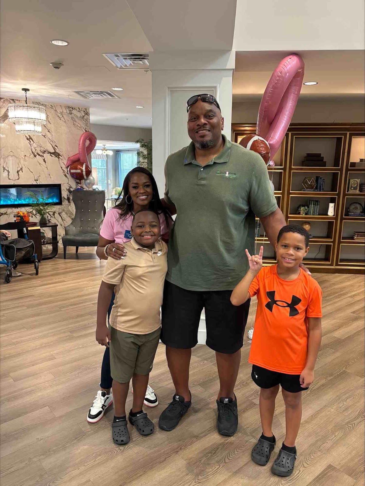 A family of four, two adults and two young boys, pose for a photo in a well-lit, modern indoor space. The father, wearing a green polo shirt and sunglasses, stands in the center. The mother, in a pink shirt, is to his left, with one son in front of her. The other son stands to the father's right, making a 'rock on' hand gesture. In the background, there are decorative elements like a fireplace, a bookshelf, and balloons that appear to be shaped like a heart and a football.