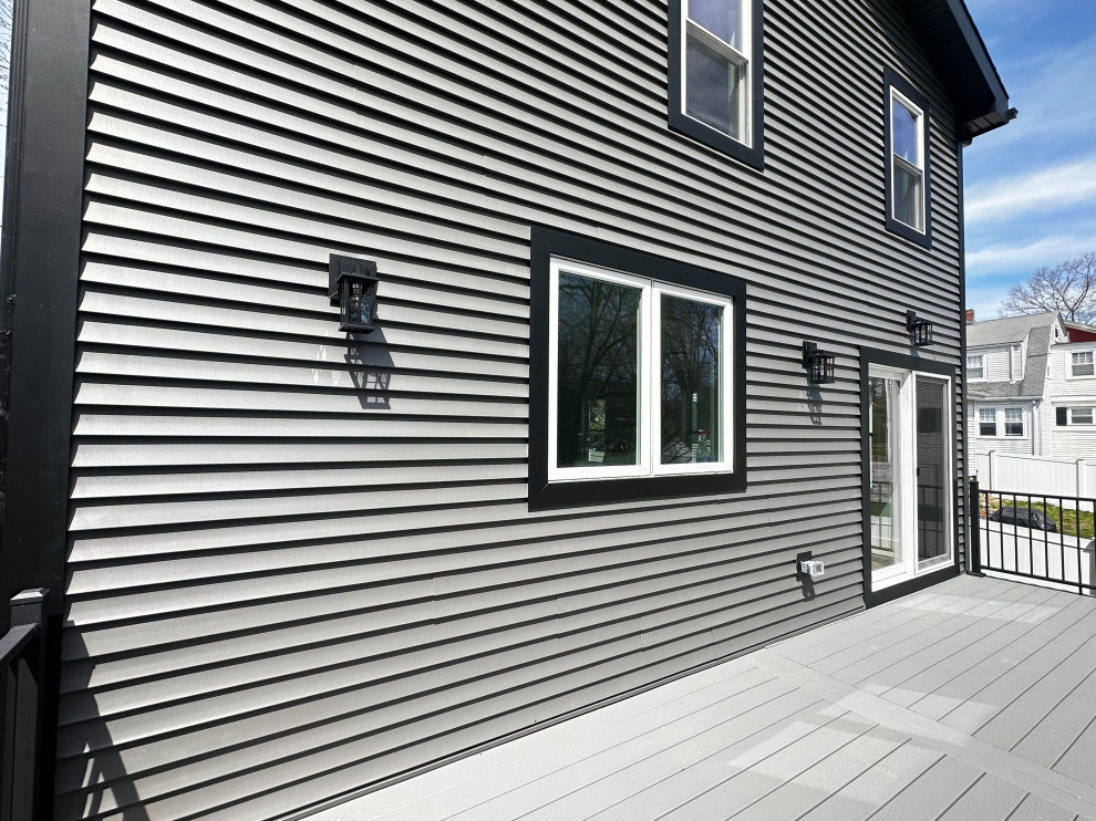 A modern house exterior with dark grey horizontal siding, black trim, and white-framed windows. A black deck with grey composite flooring extends from the house. Two black outdoor light fixtures are mounted on the siding. The sky is blue with some scattered clouds.