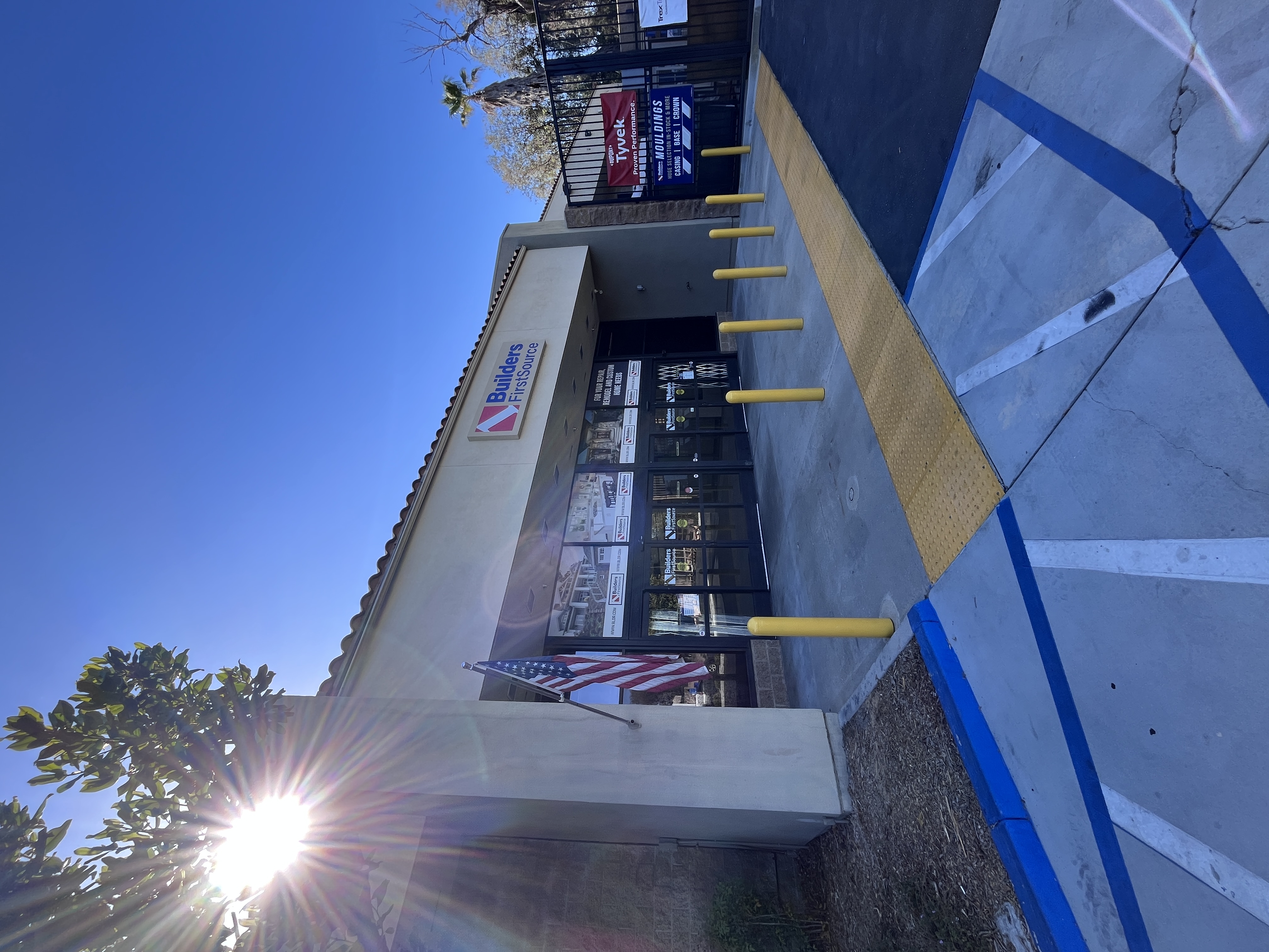 Exterior storefront entrance of Builders FirstSource Thousand Oaks, CA, with signage, an American flag, and a moulding promotion banner near the entrance.