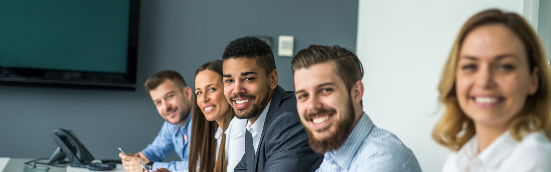 A diverse group of five professionals, four men and one woman, are seated in a line at a table, look 