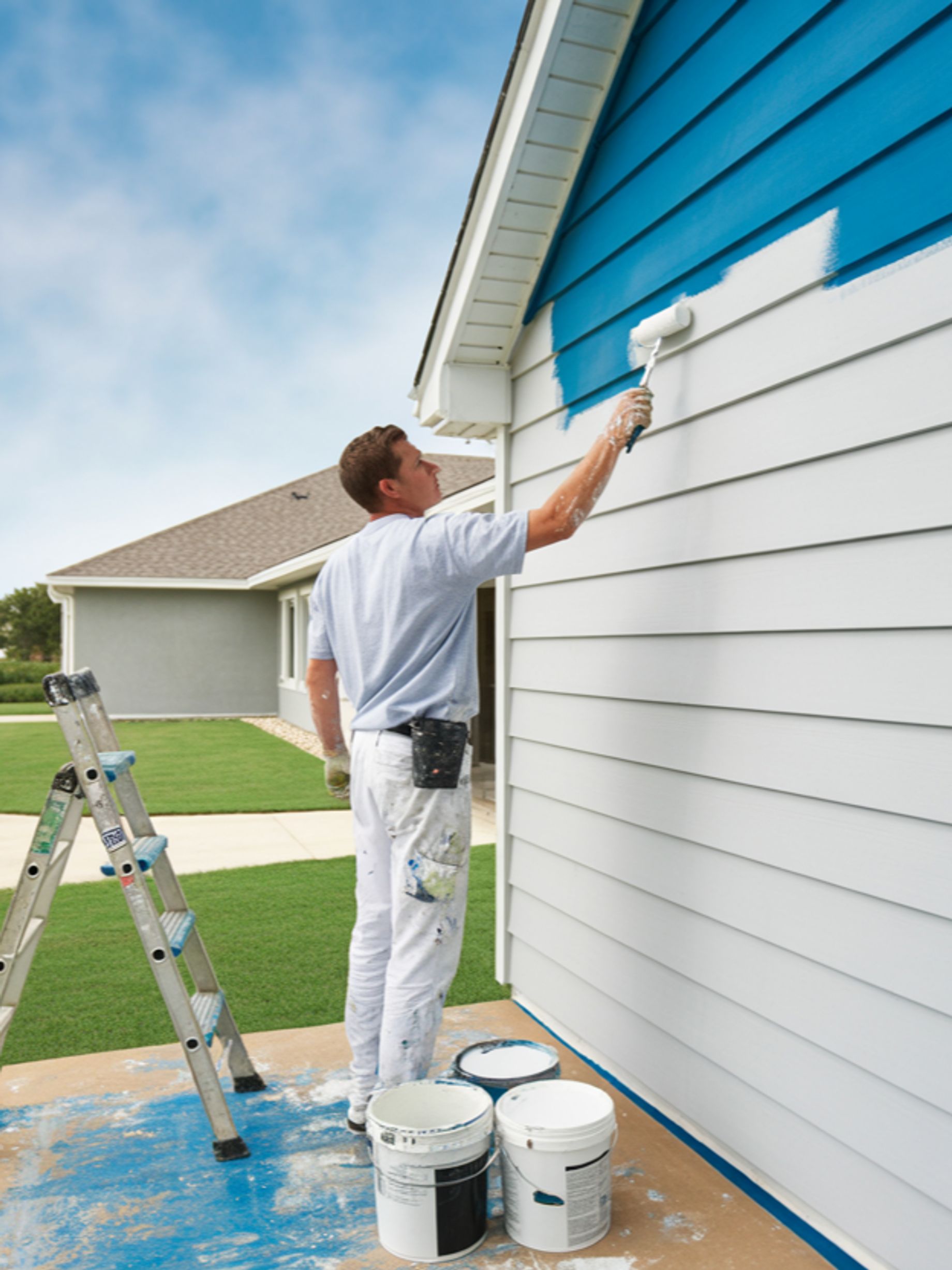 A person is painting the exterior of a house with a roller. The house has white siding and a blue ac 