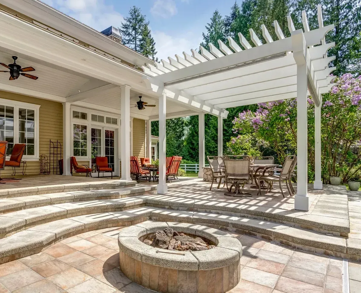 A luxurious outdoor living space featuring a tiled patio, a curved stone fire pit, and a white pergola covering an outdoor dining set. The area steps up to a covered porch with additional seating.