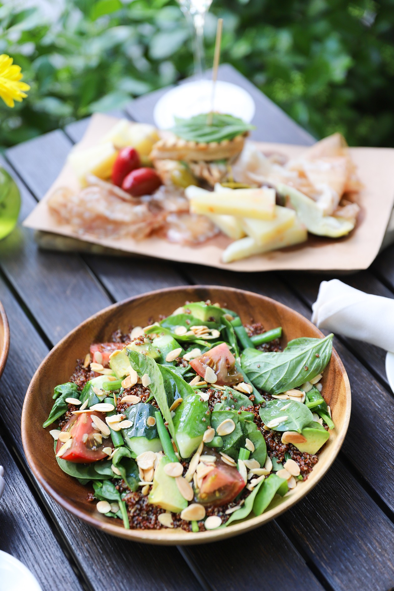 A wooden bowl filled with a quinoa salad topped with tomatoes, cucumbers, almonds, and basil. In the background, a platter of Italian cheeses, meats, and olives is served on parchment paper.