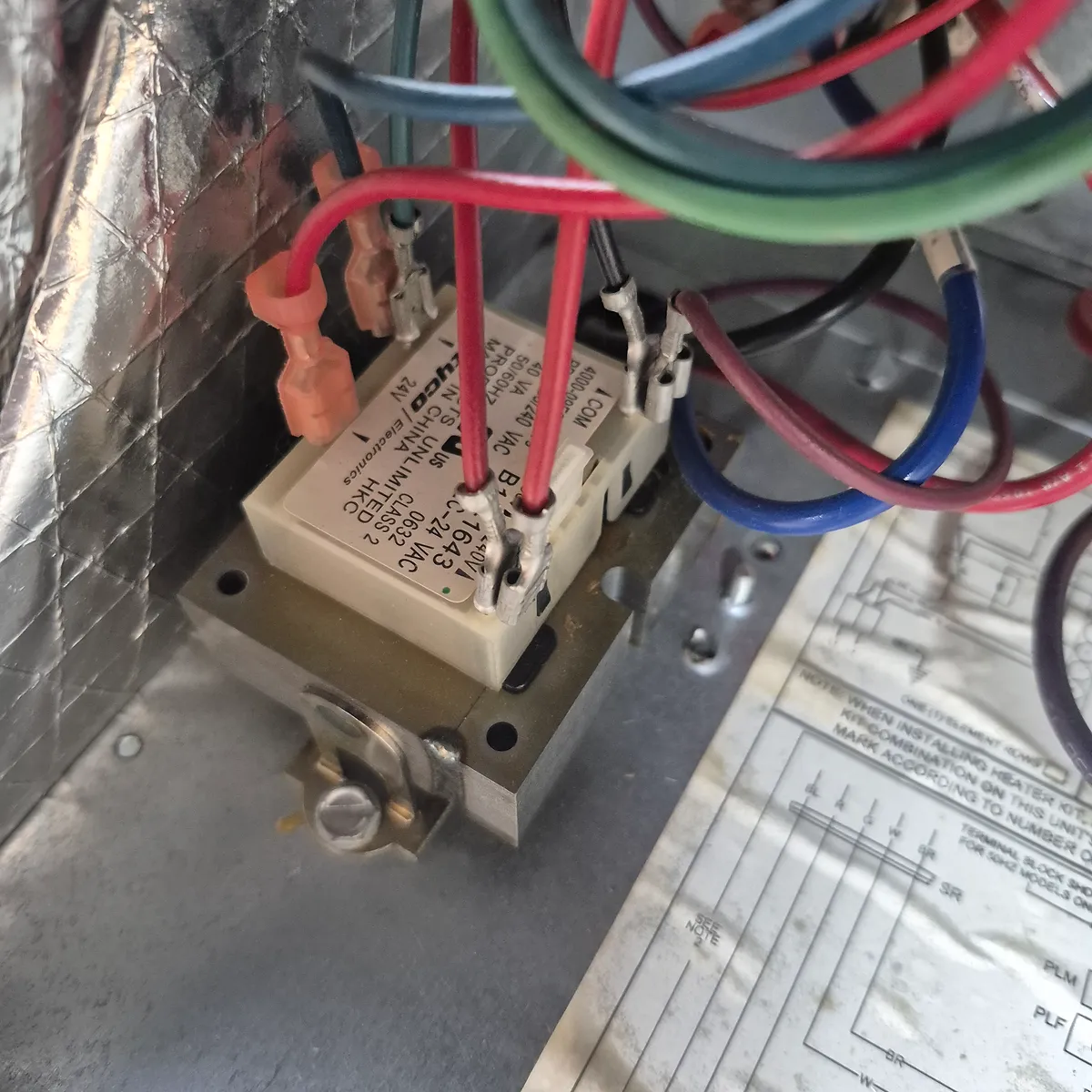 A close-up view of an electrical relay or transformer with numerous colorful wires connected to it. The component is mounted on a metal bracket within an insulated compartment. The wires are red, blue, green, and black, indicating various electrical connections. Some of the wires are connected via orange plastic connectors, while others are directly wired. A diagram or label with text is visible beneath the component.