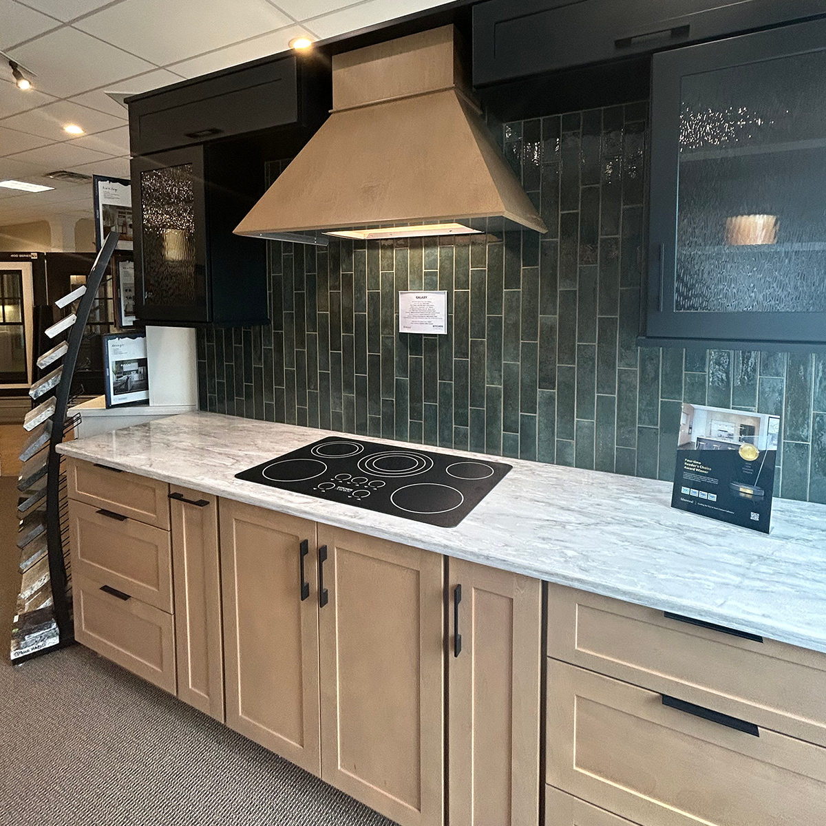 Modern kitchen showroom vignette featuring light wood cabinets, a gray marble-look countertop, black electric cooktop, green tile backsplash, and a large metal range hood surrounded by dark cabinetry.