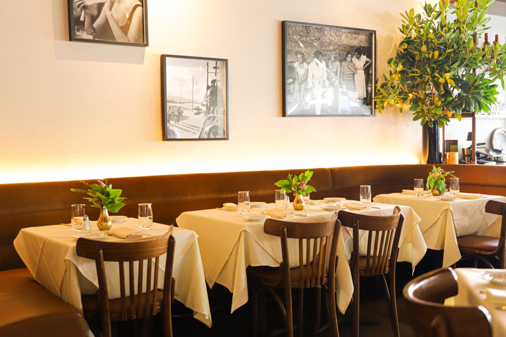 A row of elegantly set dining tables with white tablecloths, wooden chairs, and leather banquettes under soft, ambient glowing lights and framed art.