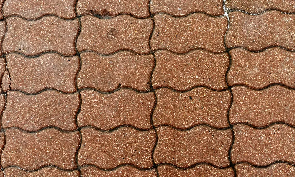 A top-down, close-up view of interlocking, wave-patterned brick pavers. The bricks are a reddish-brown color with visible aggregate, suggesting they are made of concrete or a similar composite material. The texture is rough and slightly uneven, with dark grout lines defining the spaces between the bricks. Some of the bricks appear wet, giving them a slightly darker sheen.
