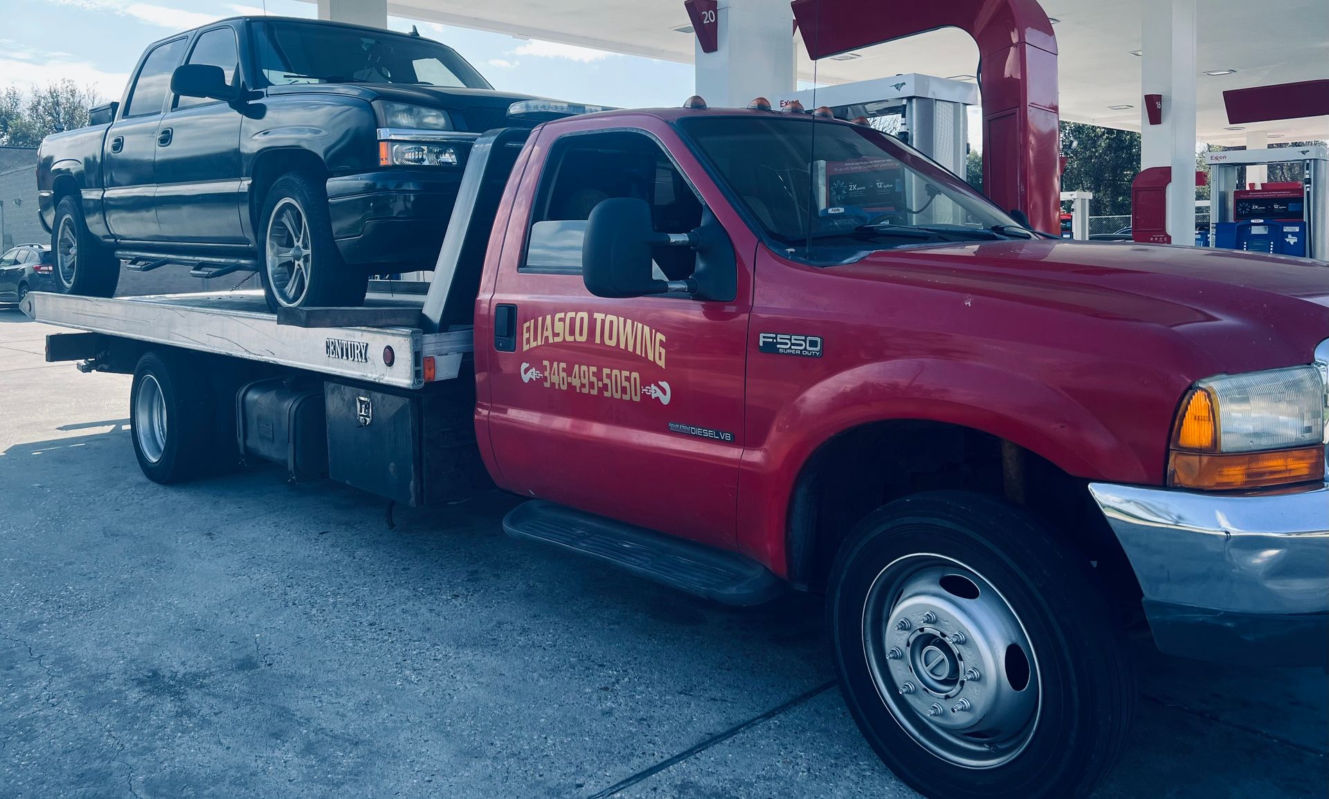 A red Eliasco Towing flatbed truck carries a dark gray Chevrolet Silverado pickup truck, positioned under the canopy of an Exxon gas station.