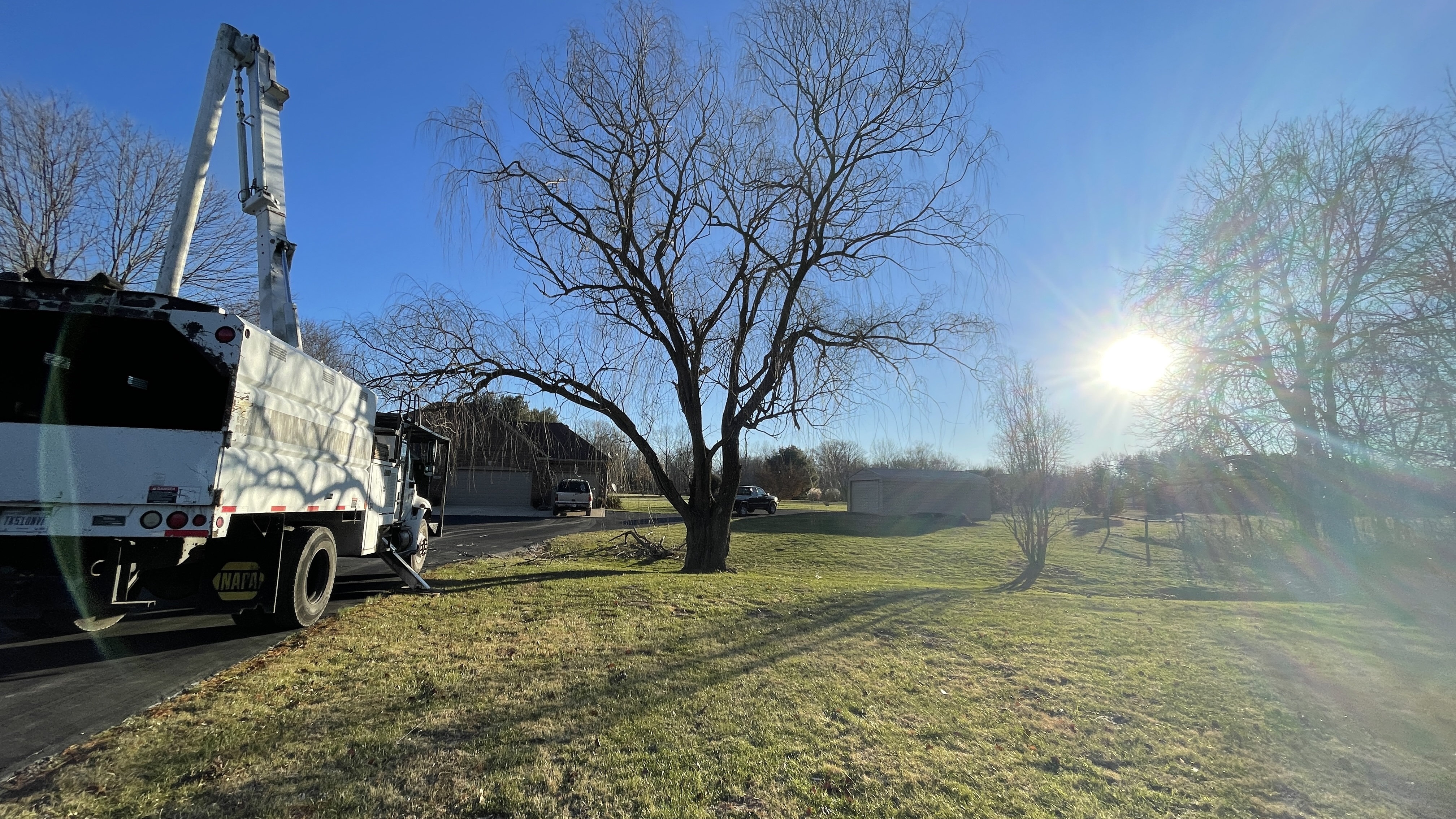 A white utility truck with a large mechanical arm is parked on a paved driveway next to a grassy lawn on a sunny day. The truck has a "NAPA" logo visible on its side. In the background, a house with a garage and a shed are visible. Bare-branched trees, including a weeping willow, dot the landscape. The sun is shining brightly, casting long shadows and creating lens flare.