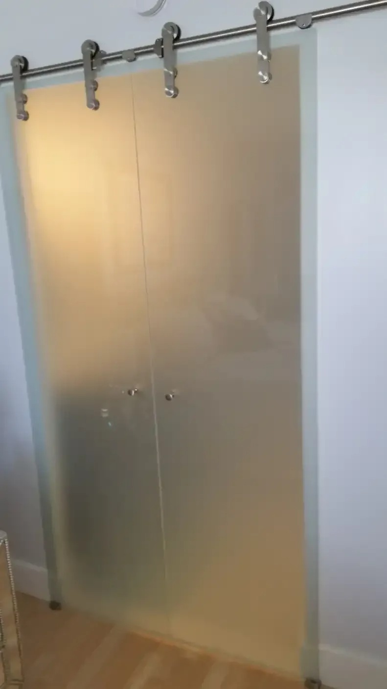 Two frosted glass panels slide on a silver track, forming a modern partition or door. The panels have small, round handles and partially obscure the view beyond.