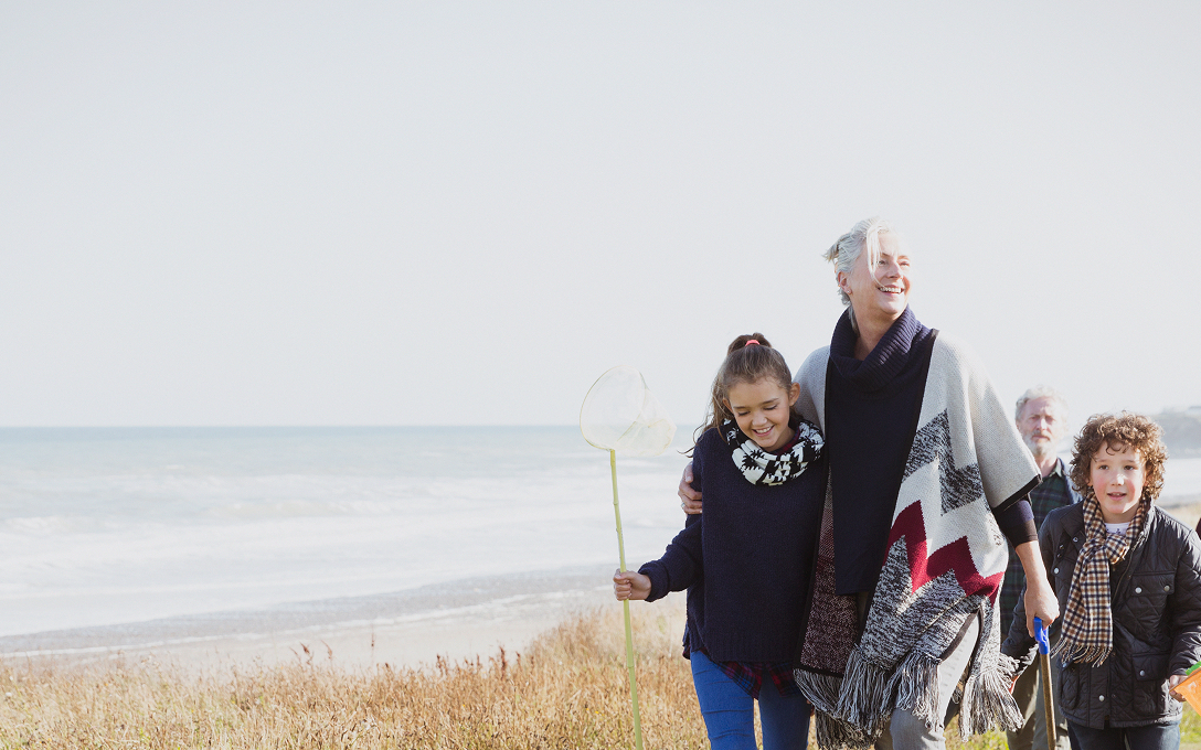 A joyful family strolls along a grassy beach path, with the ocean in the background. A young girl holds a fishing net, smiling alongside an older woman.