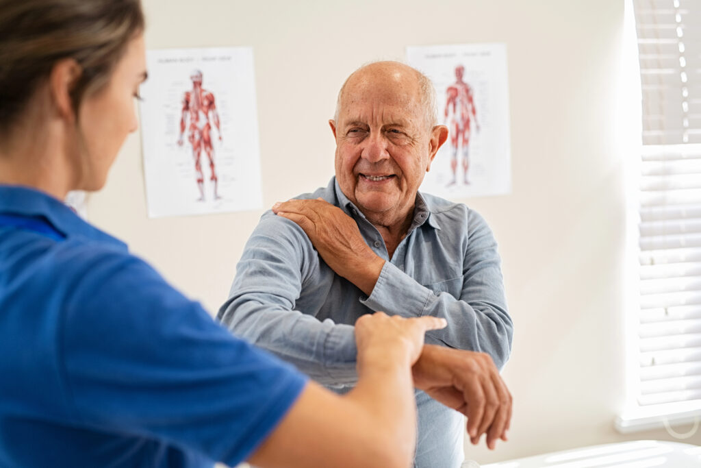 An older man with a grey shirt is being assisted by a woman in a blue uniform in a medical setting. Anatomical charts are visible on the wall in the background. The man is smiling as the woman touches his shoulder and arm.
