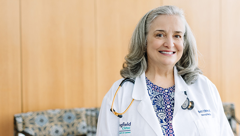 Female internal medicine doctor wearing white coat and stethoscope around her neck smiling in a patient waiting room.