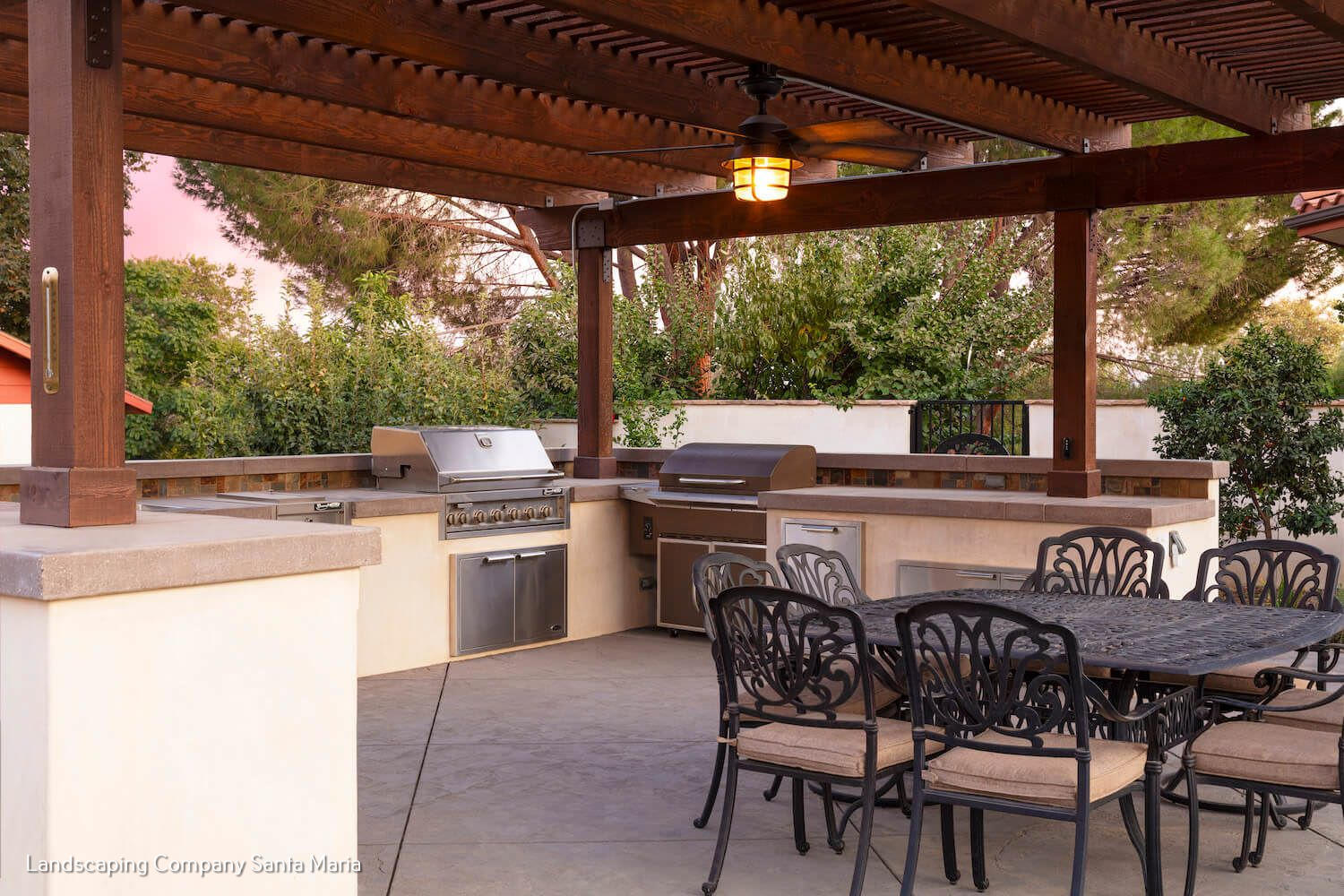 Outdoor covered patio kitchen with stainless-steel grill, bar counters, and dining table with ornate chairs. Warm lighting creates a welcoming entertaining space.