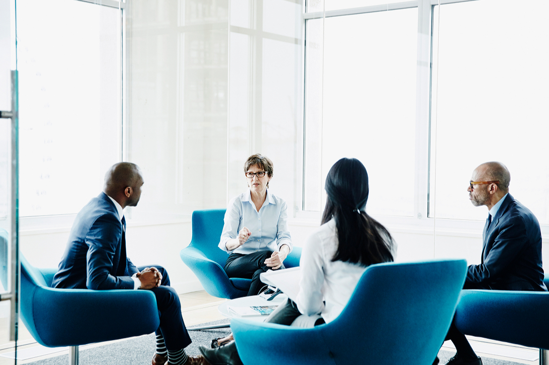 A group of four professionals sits in blue chairs in a modern office with large windows. They are engaged in a serious discussion, fostering a collaborative atmosphere.