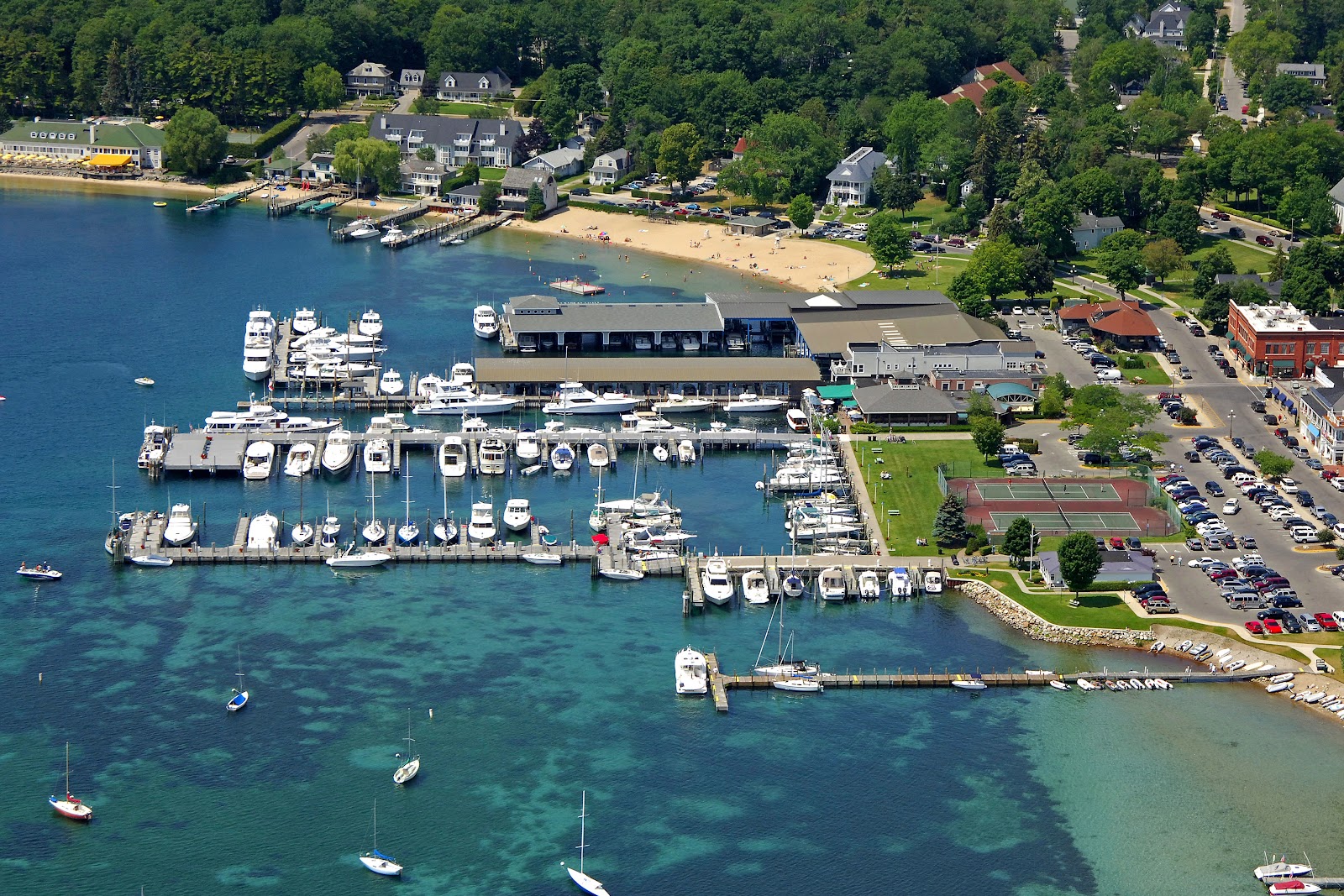 An aerial view of a bustling marina on a clear day, with numerous boats docked in turquoise waters and a lively beach scene in the background. The surrounding area features lush green trees, residential buildings, a main street with shops and parking lots, and tennis courts.