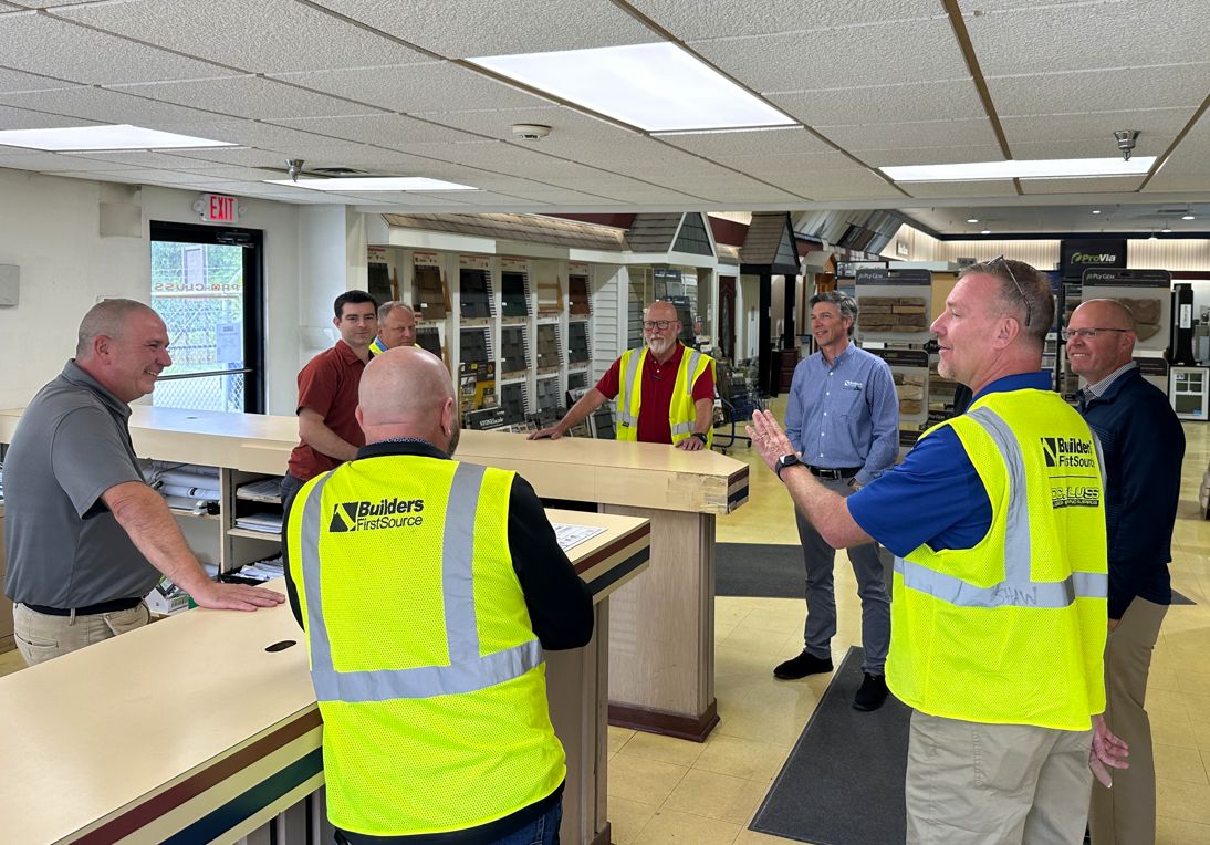 Builders FirstSource team members engage in a lively discussion at a showroom counter. Some wear yellow safety vests while others are dressed in business casual. Product displays line the background, and the group appears to be exchanging ideas or touring the facility.