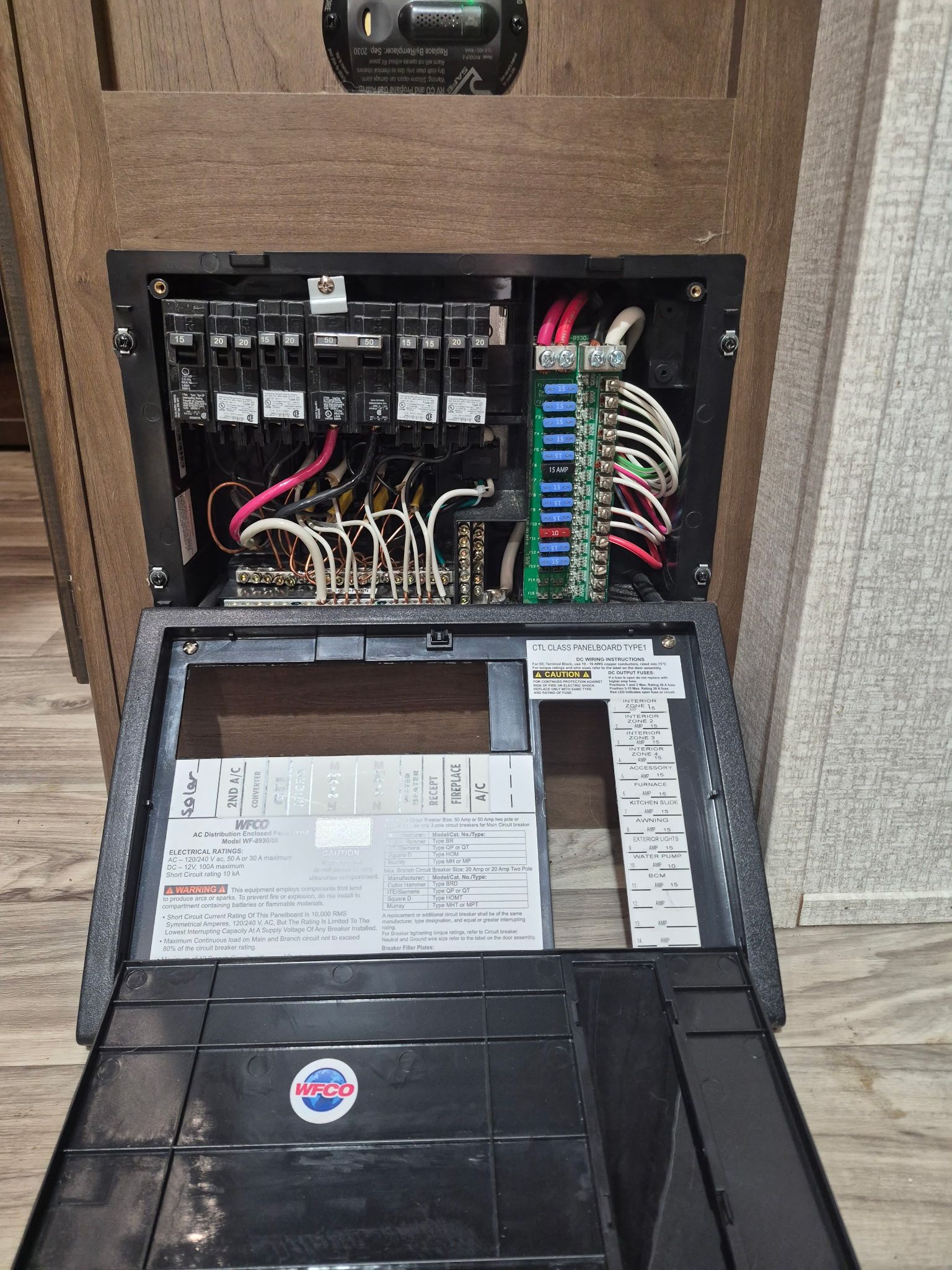 Open RV electrical breaker and fuse panel mounted in wood cabinetry, showing labeled circuit breakers, colored blade fuses, wiring terminals, distribution board, and circuit identification chart during electrical system service.