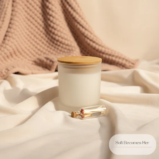 A frosted glass candle with a wooden lid sits on a bed of draped, soft, cream-colored fabric. Beside the candle is a small glass vial filled with matches. In the background, a textured, blush-pink knitted throw is draped, creating a cozy and inviting atmosphere. A text overlay reads "Soft Becomes Her".