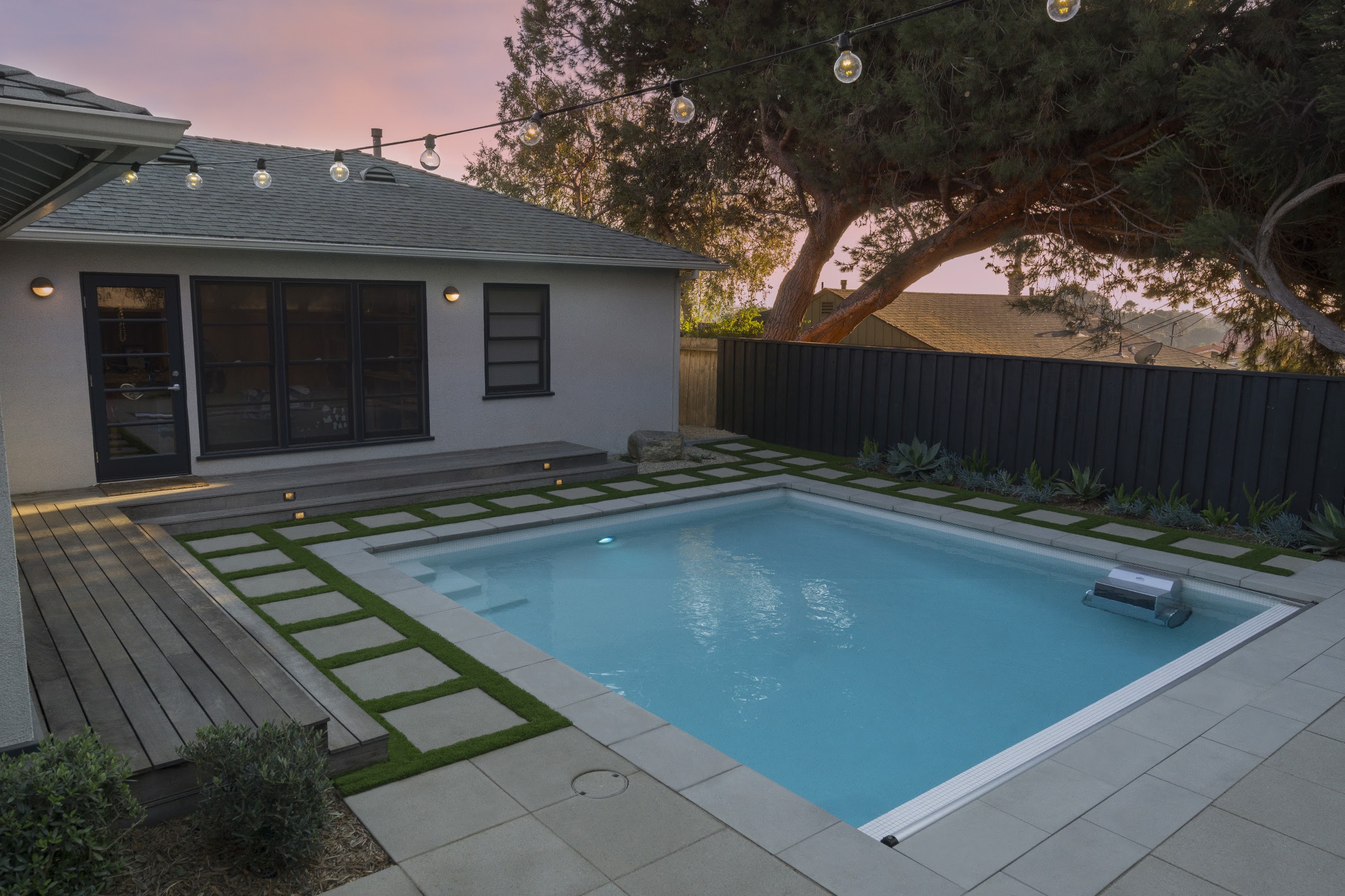 Backyard pool glows at dusk beside a modern gray home, with string lights, geometric pavers, lawn strips, trees, and a peaceful mood.