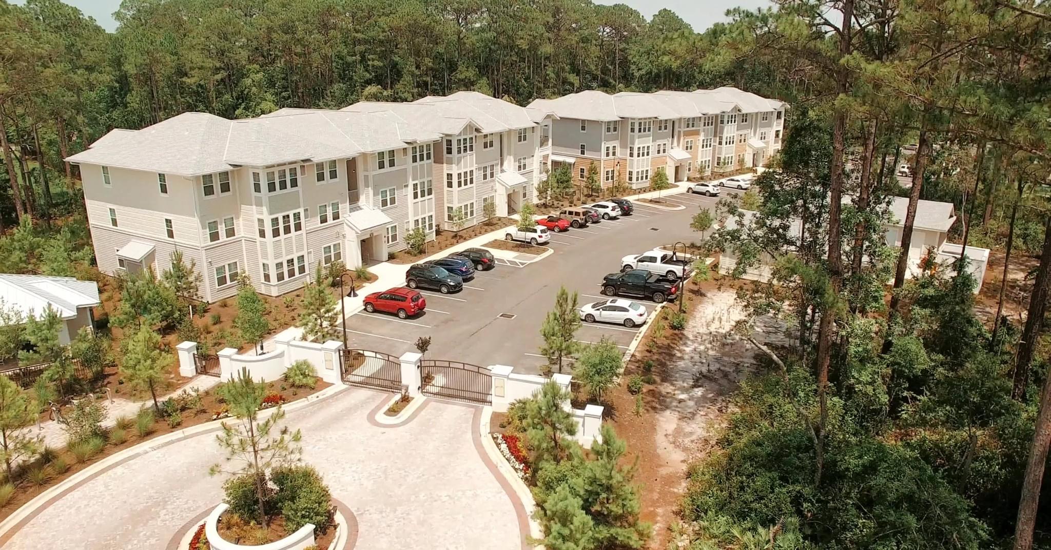 An aerial perspective shows the gated entrance and expansive parking areas of a multi-story apartment community nestled in a pine forest.