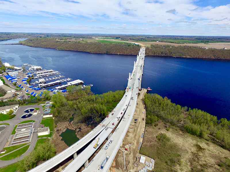 High-altitude aerial drone photograph showcasing a massive highway bridge construction project over a wide river. The multi-lane concrete bridge spans from lush green, wooded shorelines across the water, with construction equipment, staging areas, and water treatment facilities visible on the near bank.