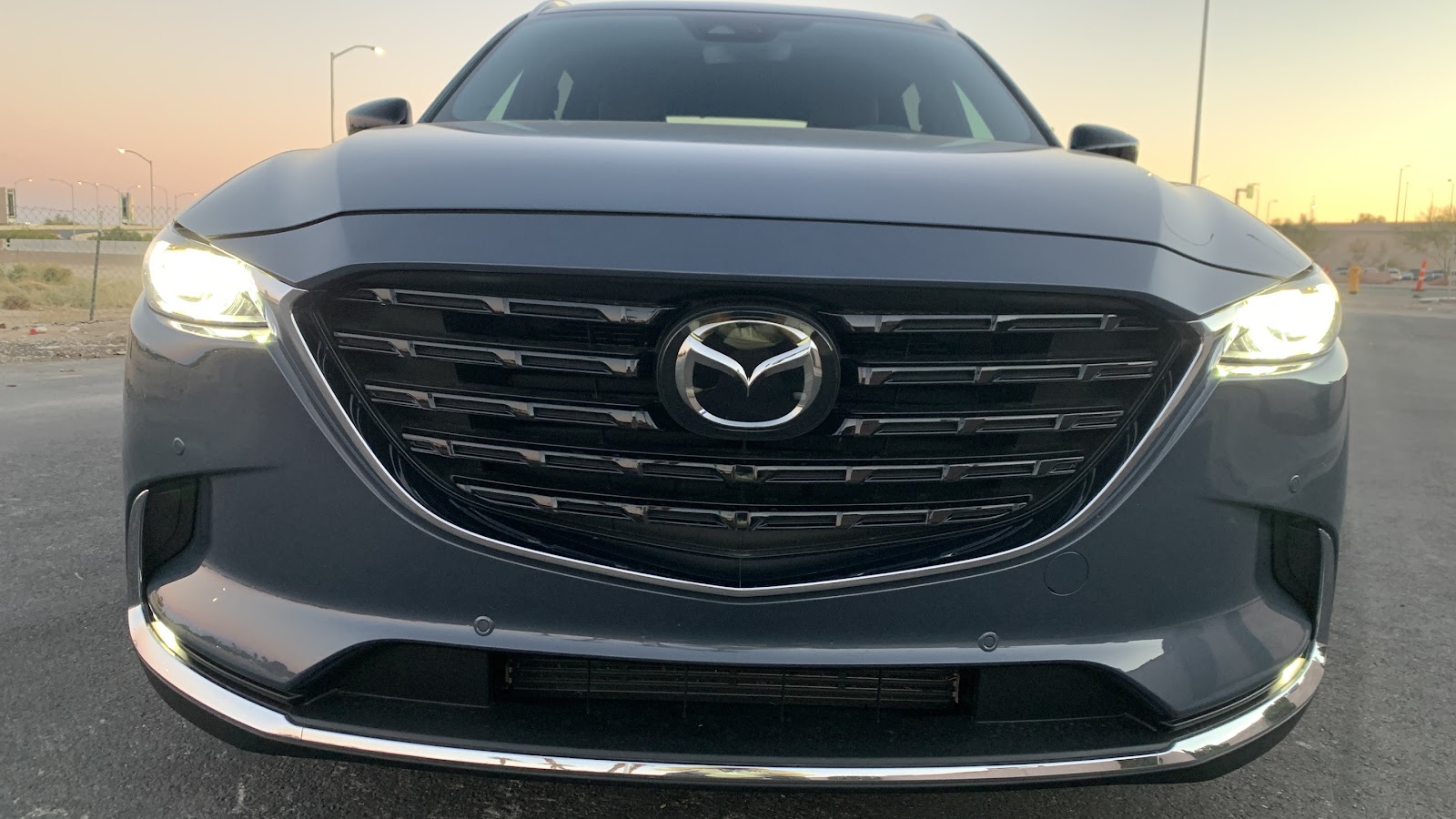 The front of a dark gray Mazda CX-9 SUV is shown up close during dusk. The car's headlights are on a 