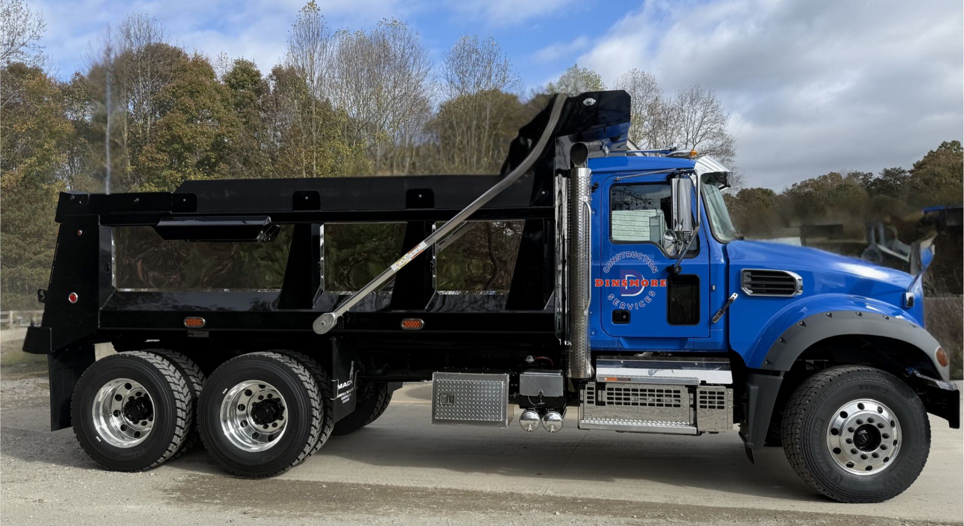 Side view of a blue Mack dump truck with Dinsmore Construction Services branding parked outdoors. The heavy duty vehicle features a large black dump bed used for hauling construction materials.