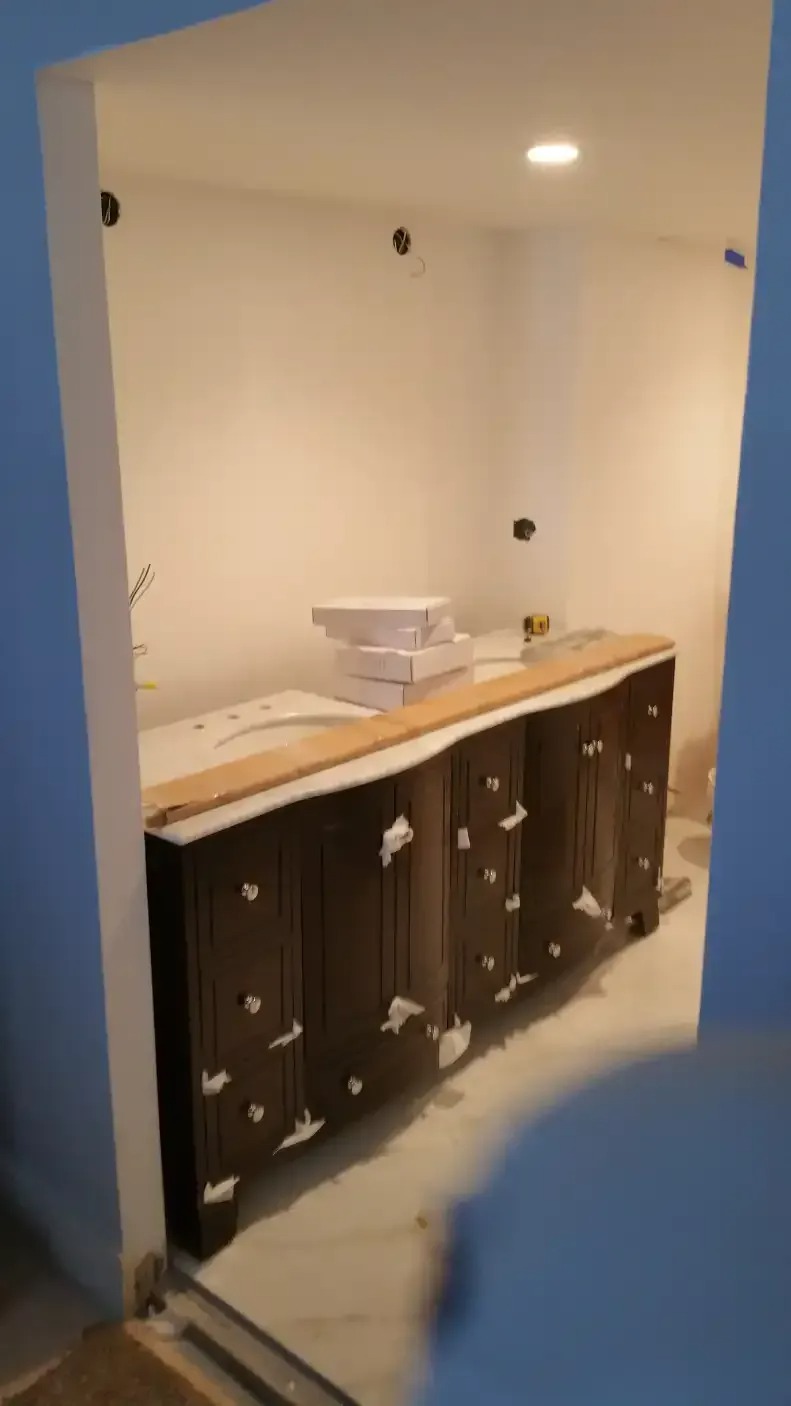A dark wood bathroom vanity with drawers is partially installed. Boxes are stacked on the countertop, and the floor is unfinished. The room is under construction, with exposed walls.