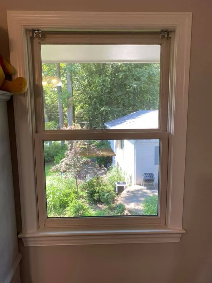 A view through a white double-hung window showing a lush green backyard with trees, bushes, a small patio with a table and chairs, and a white house. A stuffed toy bear is visible on the left side of the window frame inside the room.