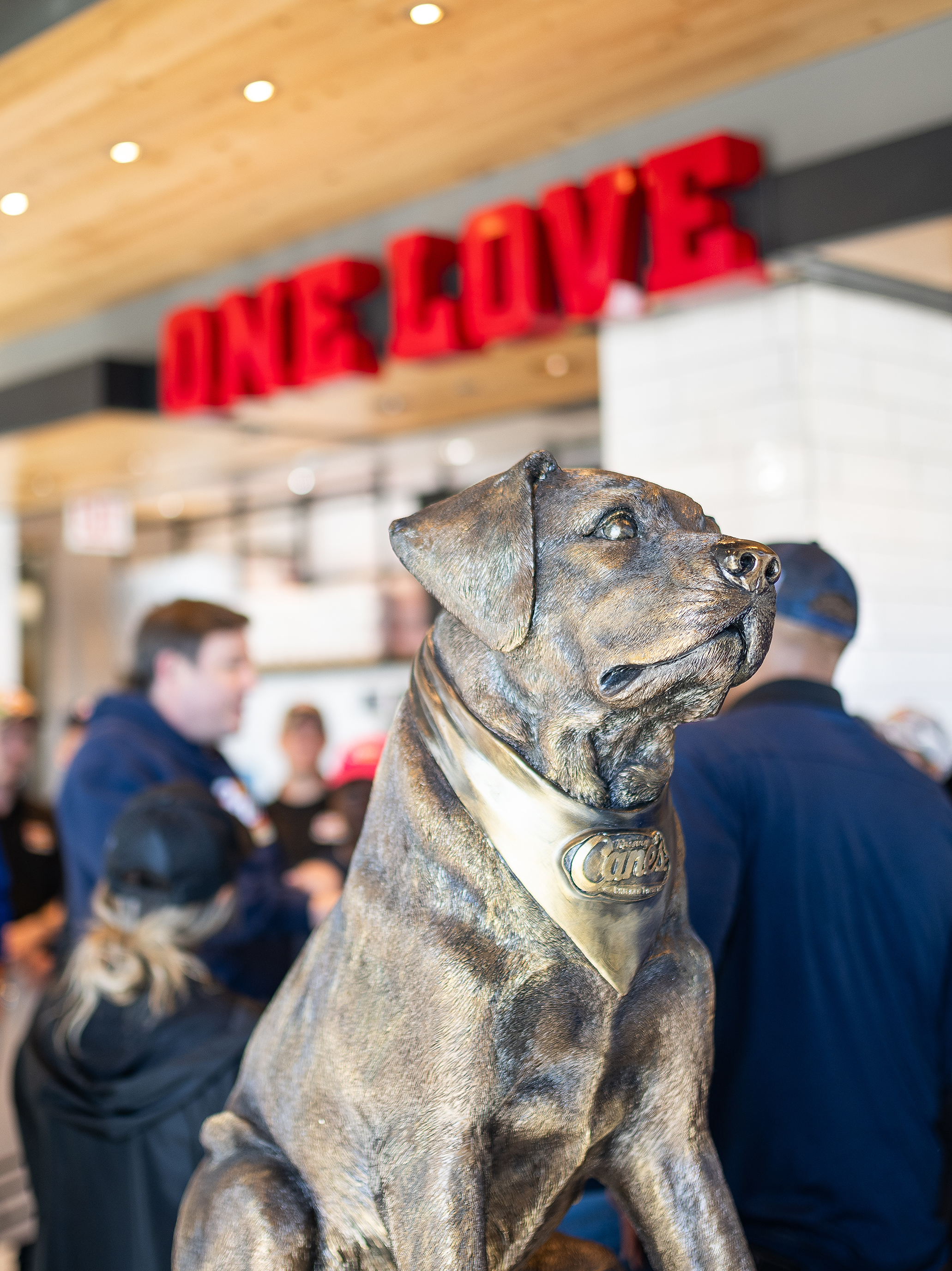 A close-up shot of a bronze statue depicting the Raising Cane's dog mascot, sitting attentively and wearing a bandana engraved with the Raising Cane's logo. In the blurred background, there are people in a busy restaurant setting underneath a bright red neon "ONE LOVE" sign.