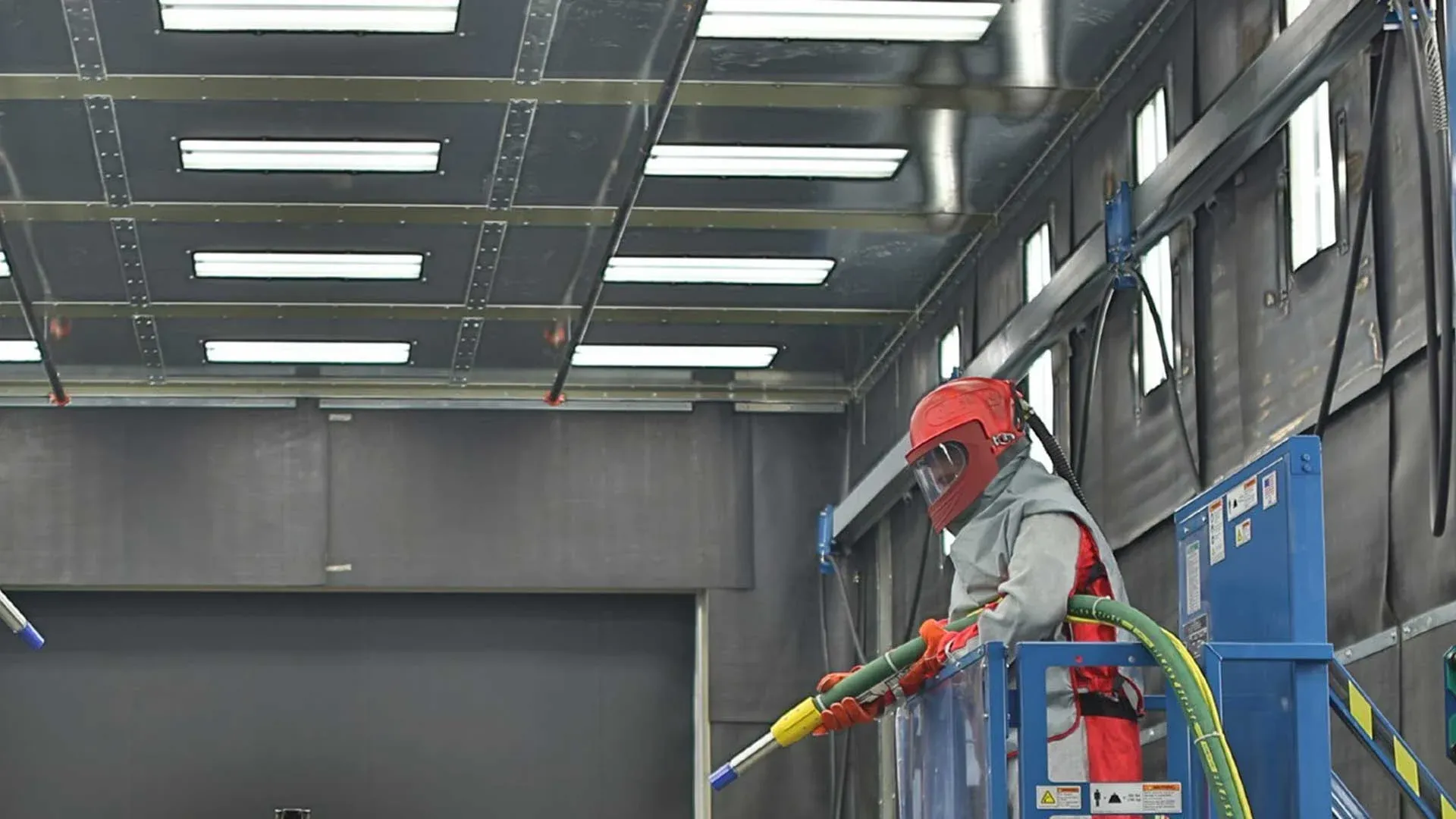 Technician wearing full protective gear operates industrial sandblasting equipment inside a controlled facility, preparing metal surfaces for coating, ensuring precision cleaning, surface profiling, and high-quality finishing standards.