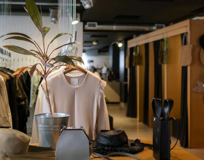 An interior view of a high-end clothing boutique showing racks of neutral-toned apparel, potted decorative plants, handbag accessories on display counters, and changing rooms in the background.