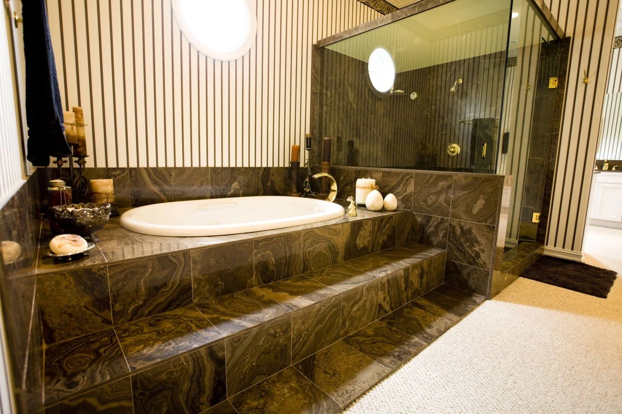 A luxurious bathroom features a sunken oval bathtub surrounded by dark, marbled tile steps. A separate glass-enclosed shower is visible in the background. The walls are adorned with vertical striped wallpaper and a small circular window. Decorative candles, soap, and two white decorative eggs are placed on the edge of the bathtub.