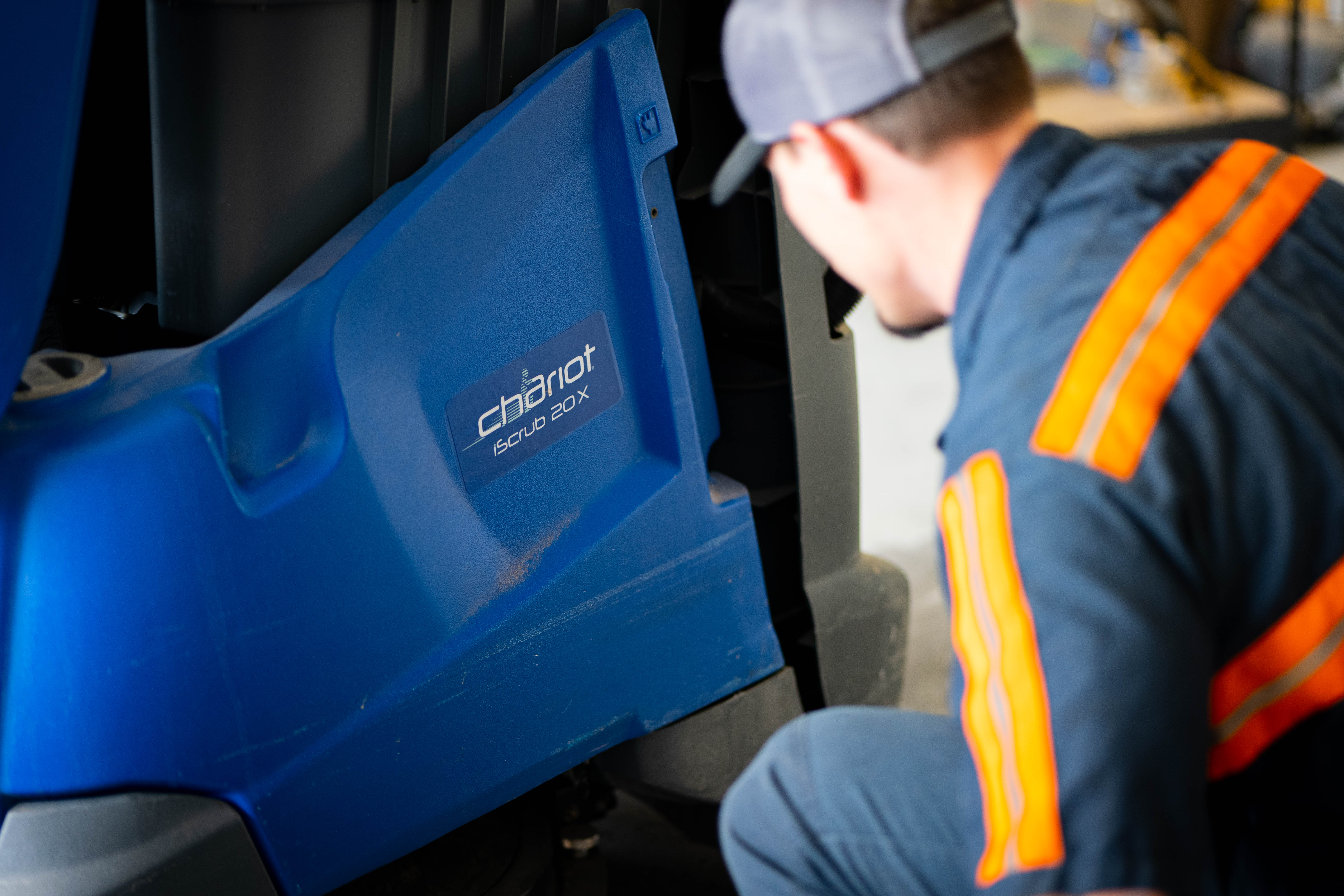 A professional WaySolutions field technician performs a meticulous visual inspection on the lower chassis of a blue Kärcher Windsor Chariot iScrub 20X stand-on floor scrubber. Serving Layton, Utah, our experts thoroughly assess exterior wear and structural integrity before beginning deep mechanical repairs to ensure maximum uptime for commercial cleaning fleets.
