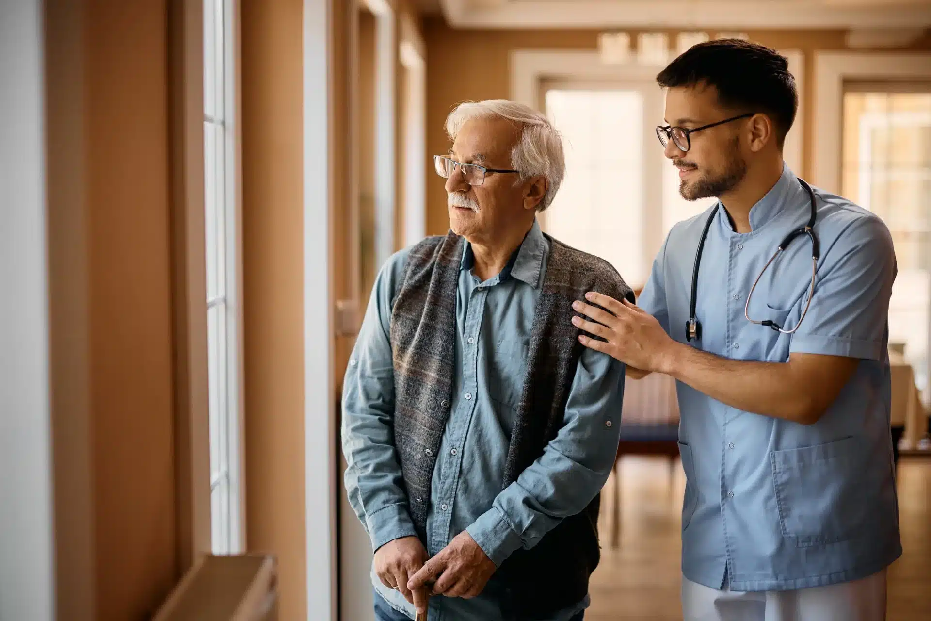 A male caregiver in a light blue uniform with a stethoscope around his neck places a reassuring hand on the shoulder of an elderly man. The senior man, wearing glasses and a vest over a blue shirt, holds a cane and looks out a window. They are in a well-lit hallway that appears to be part of a nursing home or assisted living facility.