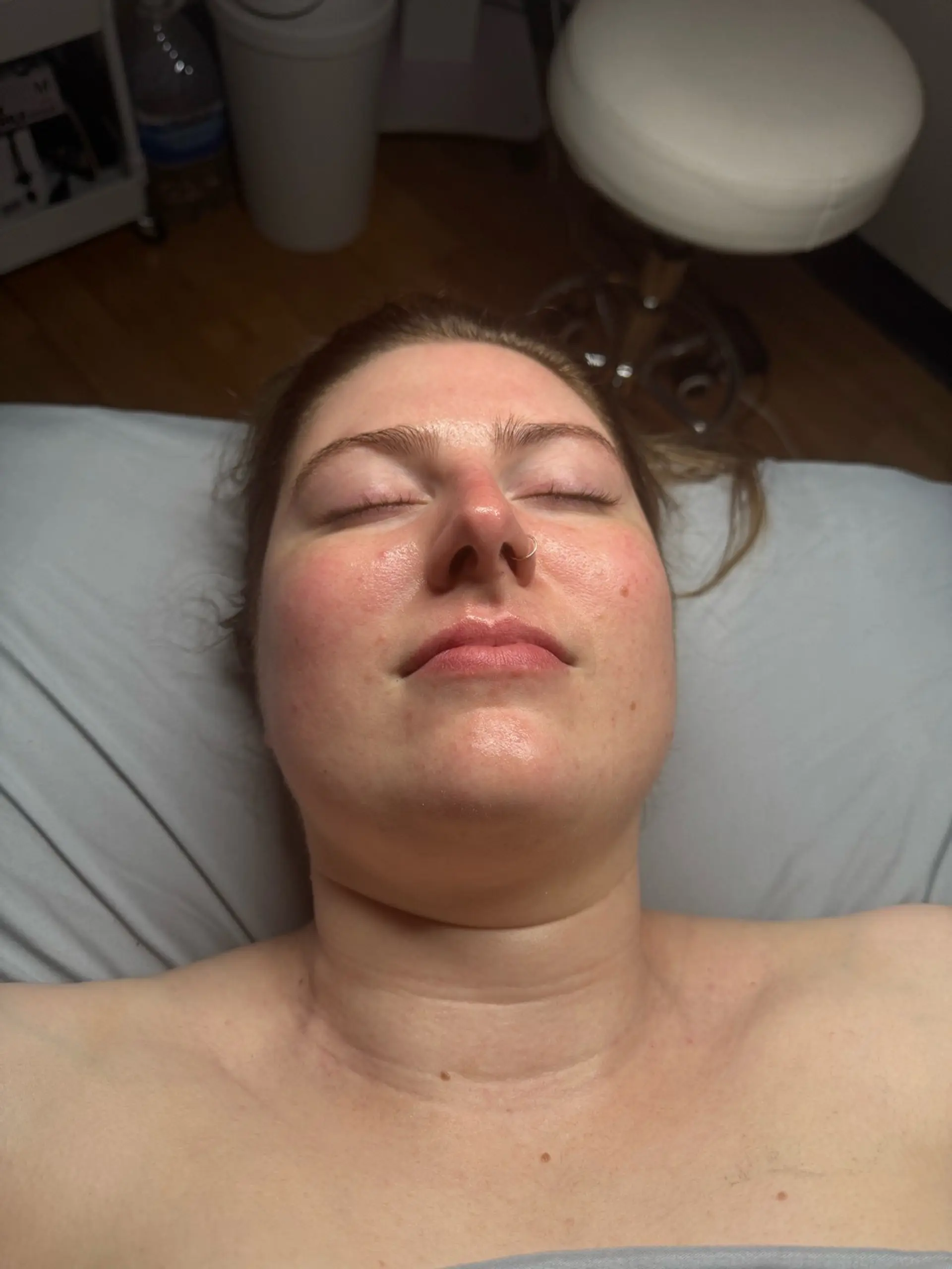 A close-up, overhead shot of a woman's face with her eyes closed, lying on a grey and white striped surface. She has a gold hoop nose ring and fair skin with some redness on her cheeks. The background shows a wooden floor, a white rolling cart with items on it, a clear plastic bottle of water, and a white rolling stool.