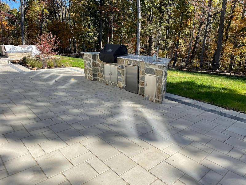 Outdoor patio featuring built-in stone kitchen and grill area surrounded by paved surface. The design blends functionality and style, creating a perfect space for outdoor cooking and entertaining with durable materials and professional landscape construction.