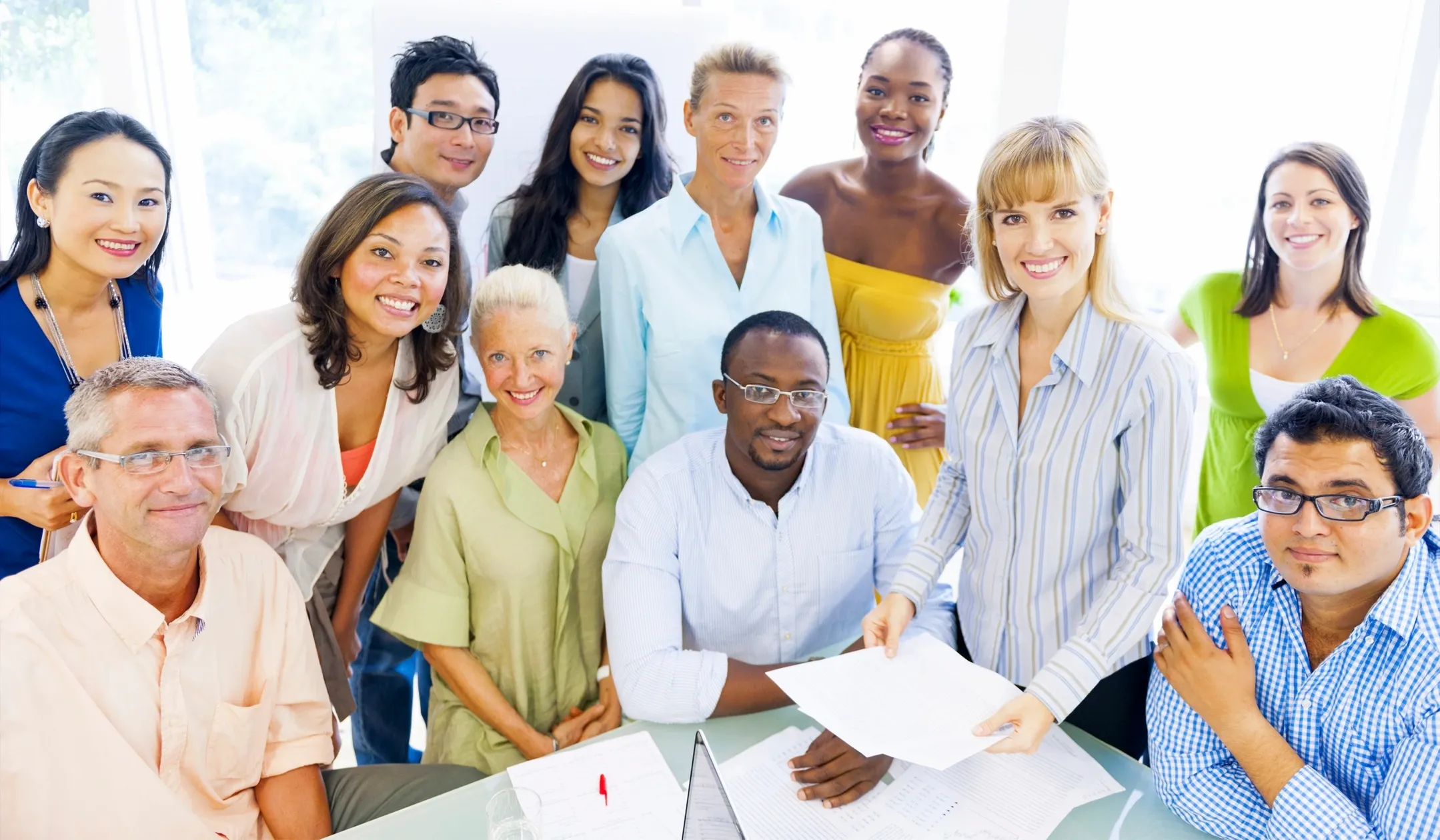 A diverse group of ten adults, appearing to be colleagues, are gathered in a bright, well-lit room, possibly an office or meeting space. They are arranged in a casual, semi-circle formation, with some standing and others seated at a table. Most of the individuals are smiling and looking towards the camera, suggesting a posed photograph. The overall atmosphere is friendly and professional. The people are dressed in business casual attire, including shirts, blouses, and dresses in various colors. Some are holding documents or interacting with papers on the table, indicating a work-related context. The background is out of focus, but suggests windows with natural light and possibly some greenery outside. The group comprises individuals of different ethnicities, ages, and genders, representing a multicultural workforce.