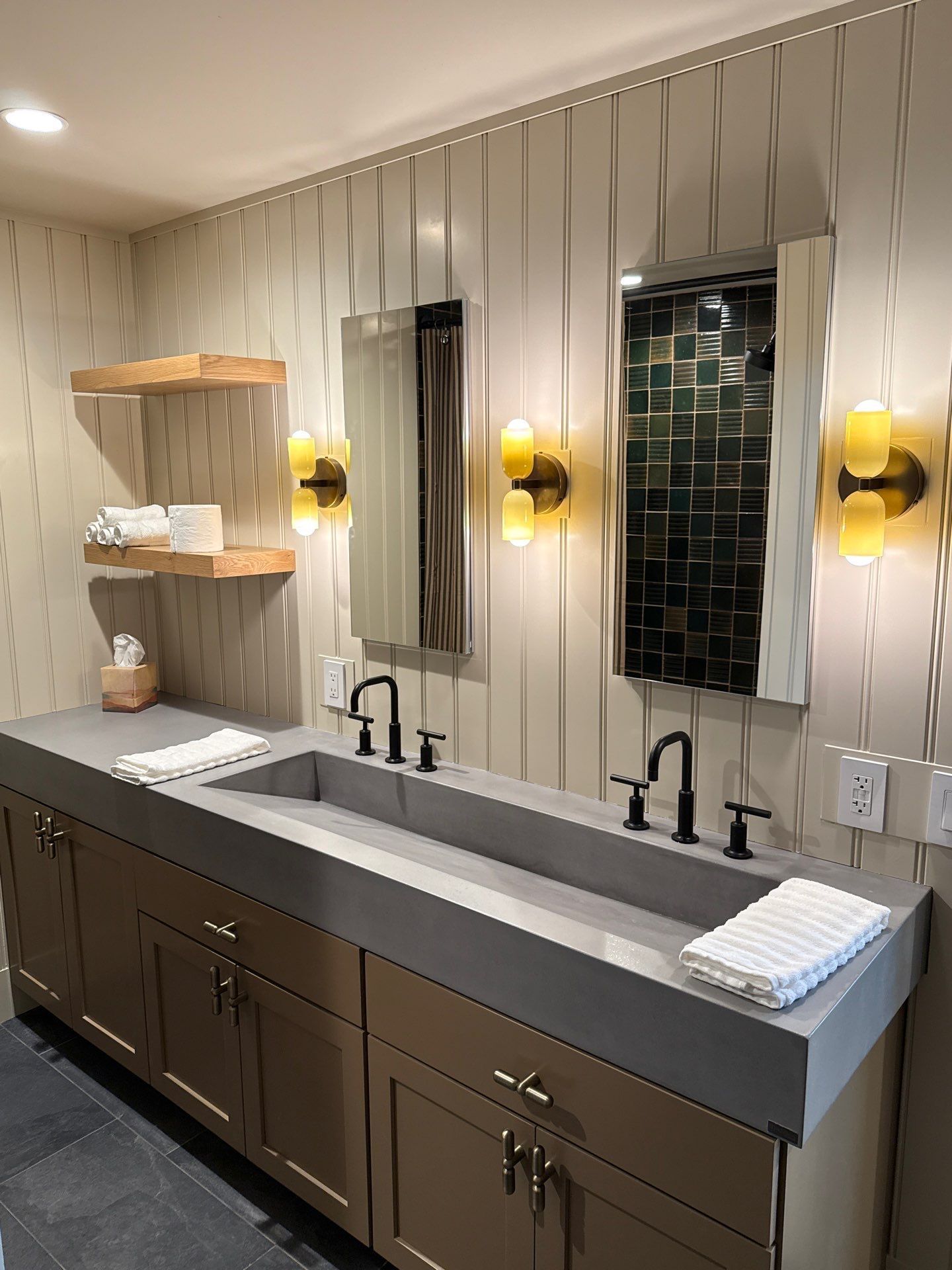 Modern double vanity bathroom features a long concrete countertop, two integrated sinks, black faucets, tall rectangular mirrors, warm wall sconces, brown cabinetry, floating wooden shelves, and folded white bath towels.