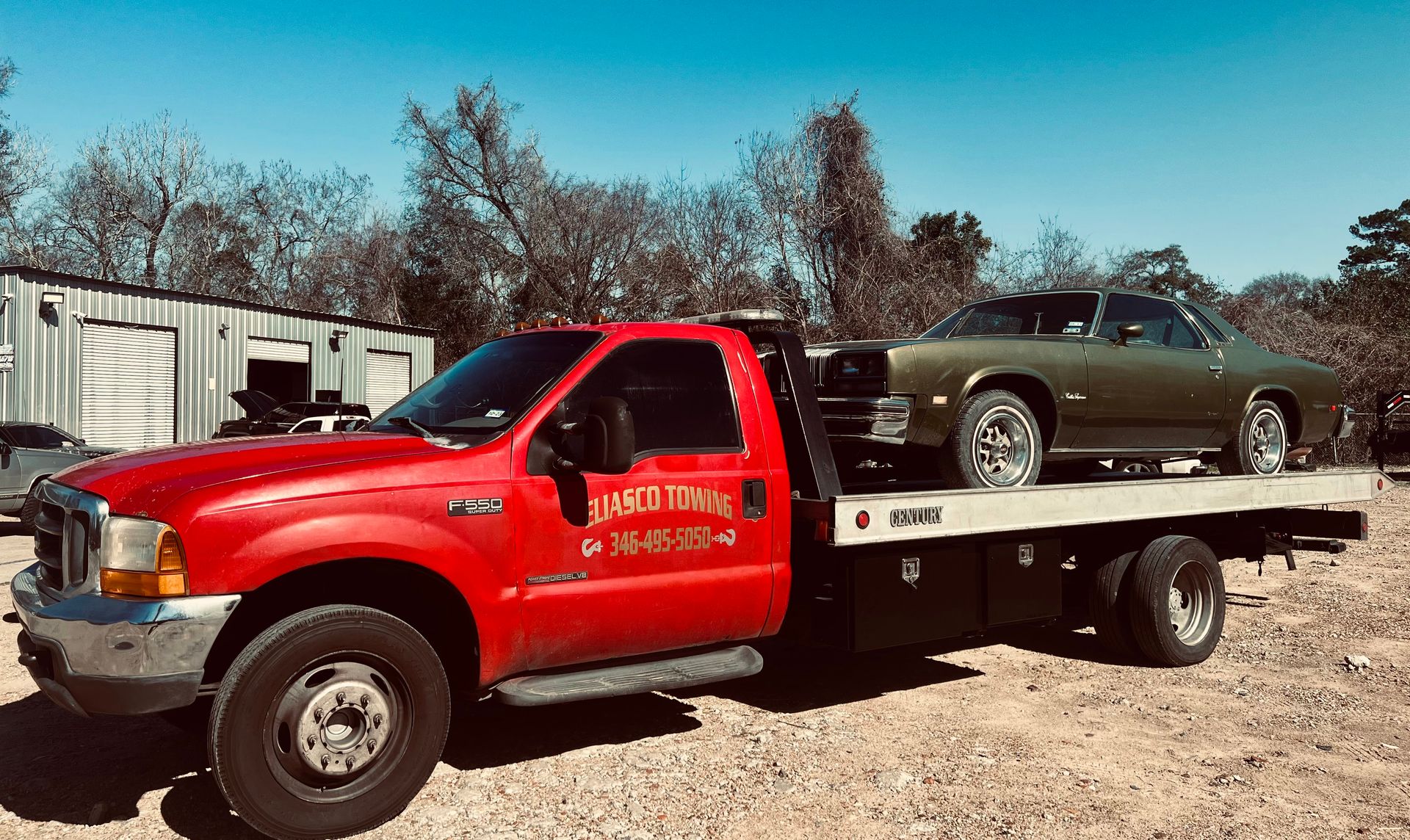 A red Eliasco Towing F-550 flatbed truck hauls a vintage olive-green two-door coupe, parked outside a metal garage building surrounded by bare winter trees.