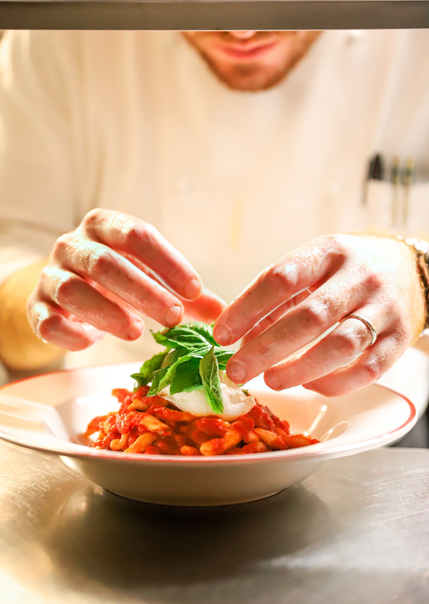 A chef in a white uniform carefully places fresh green basil leaves atop a plate of pasta with red sauce and creamy cheese. The close-up shot highlights the texture of the food.